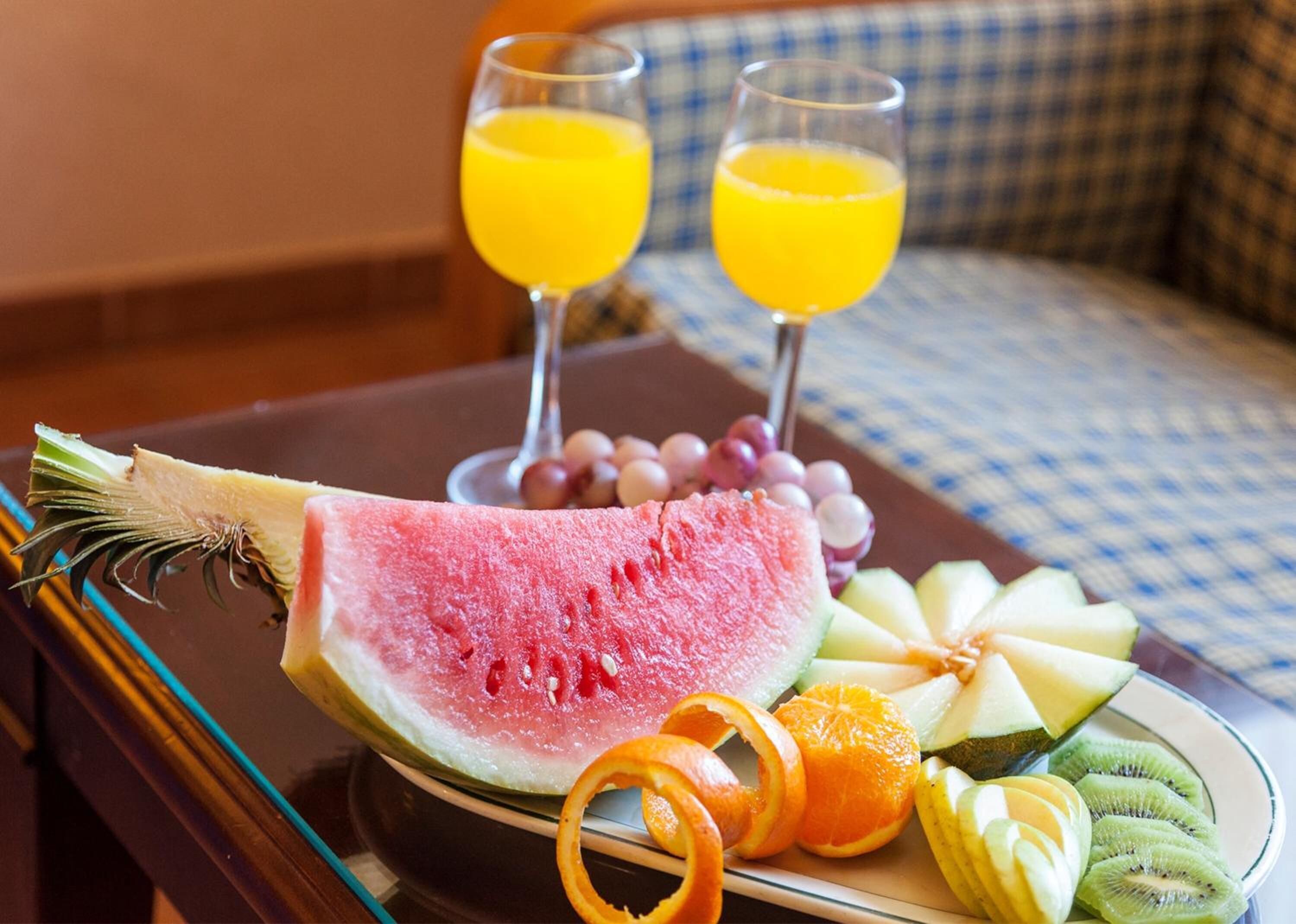 Una bandeja con una rebanada de sandía, kiwi, rodajas de naranja, melón, uvas y dos vasos de jugo de naranja sobre un mantel a cuadros.