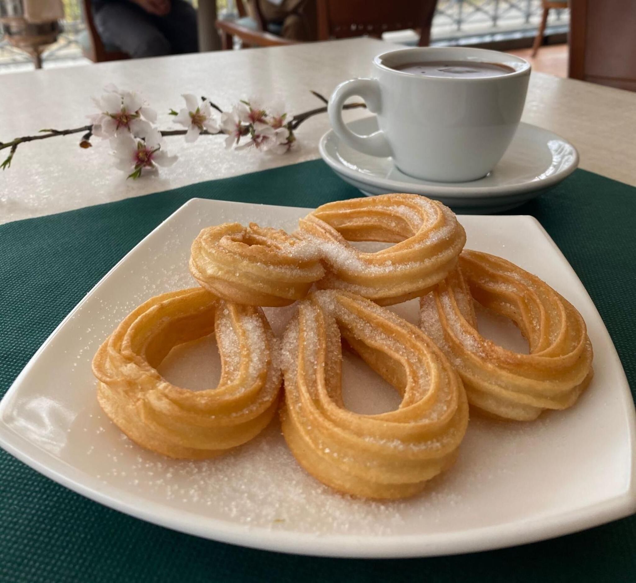 A plate of churros with sugar on a white plate, a cup of coffee, and a green tablecloth.