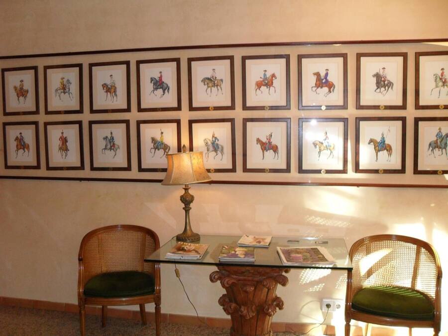 Une pièce comprenant une table en verre, deux chaises en osier, une lampe et un mur orné d’œuvres d’art équestres encadrées.