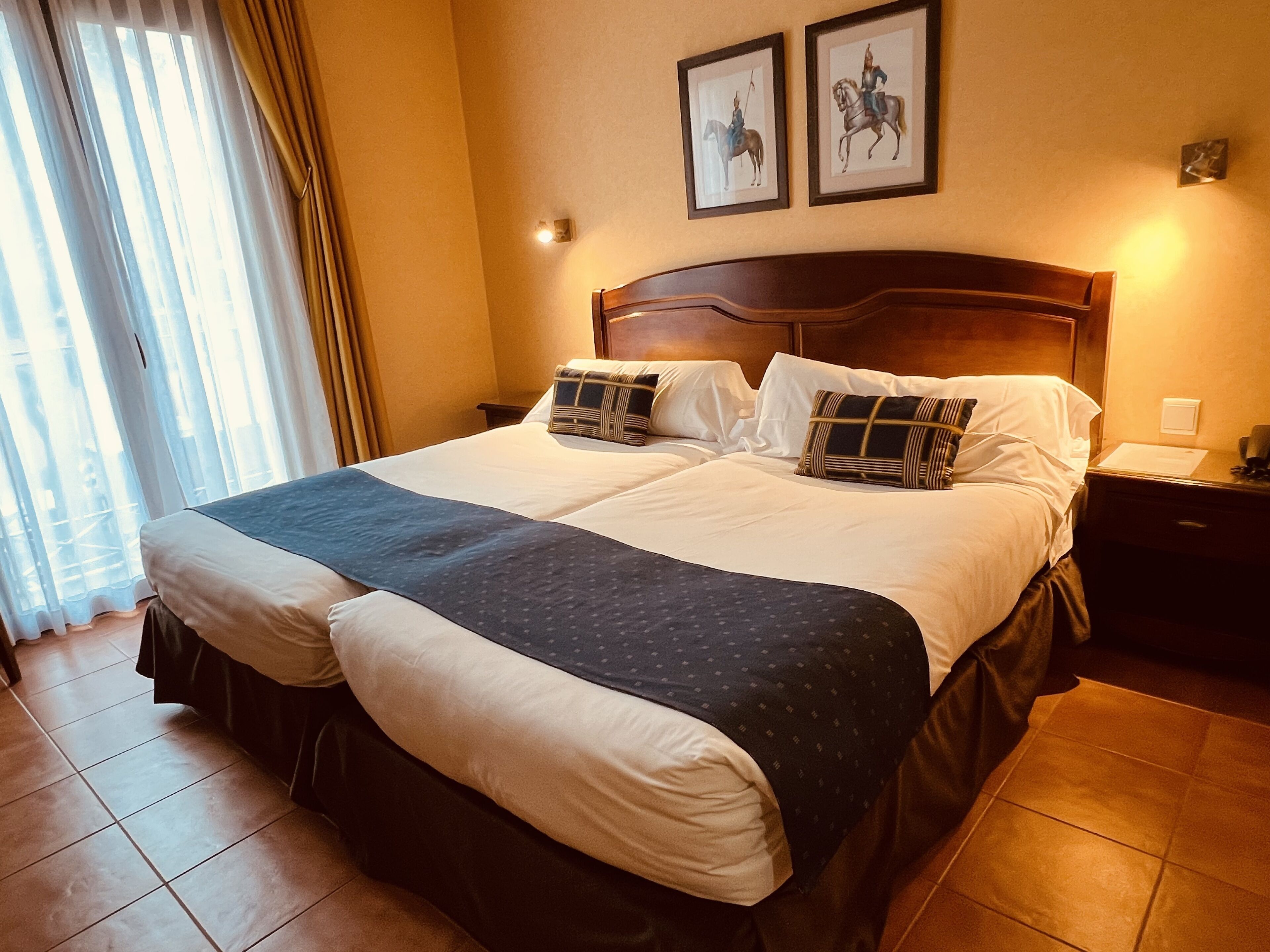 A hotel room with a large bed, two framed pictures on the wall, a nightstand, and a window with curtains.