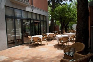 An outdoor patio area with tables and wicker chairs, surrounded by trees and a building with large glass doors. - Hospedium Hotel Los Lanceros (San Lorenzo de El Escorial)