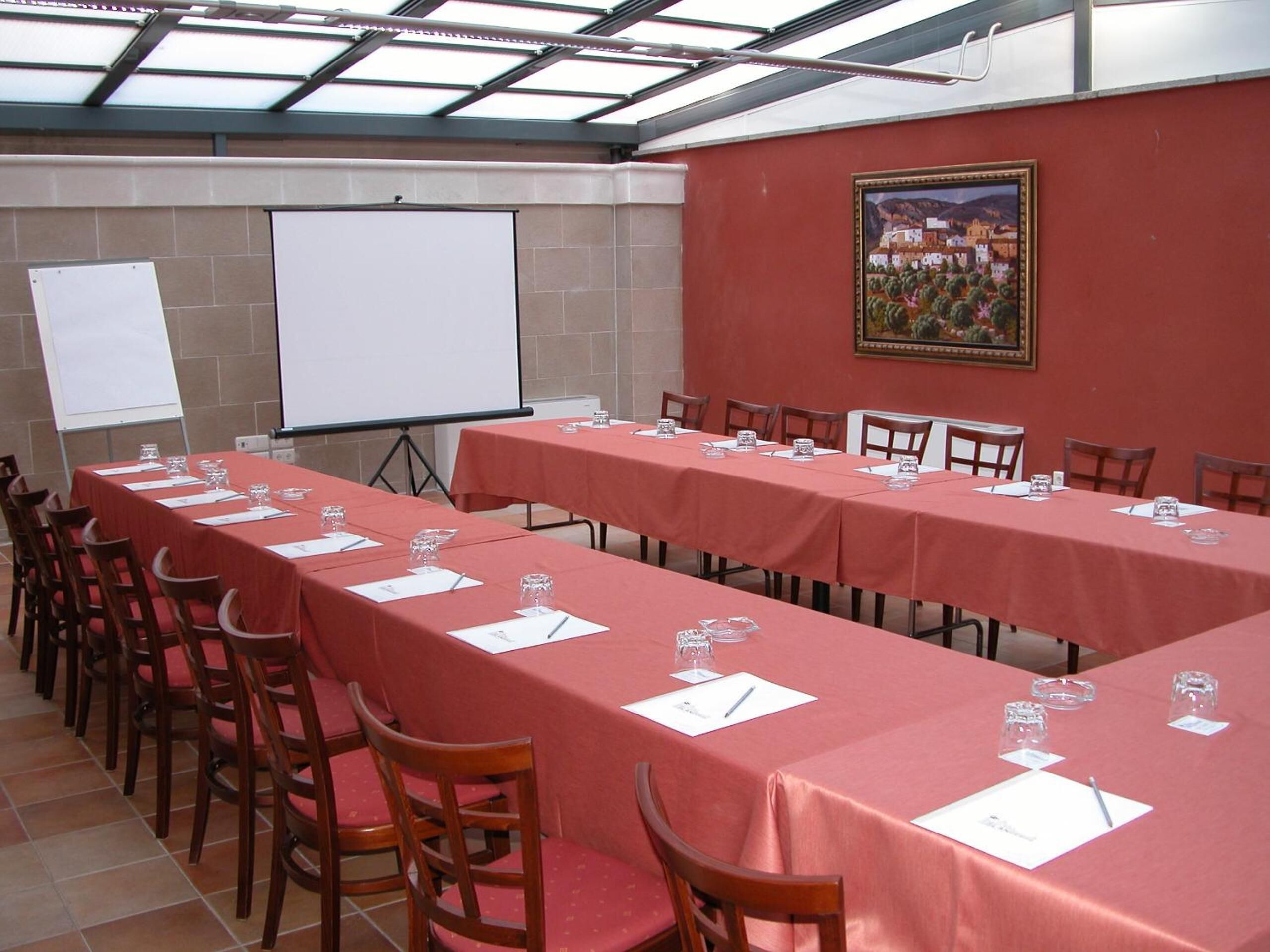 Un salón de conferencias con mesas largas, sillas, una pizarra y una pantalla de proyector.
