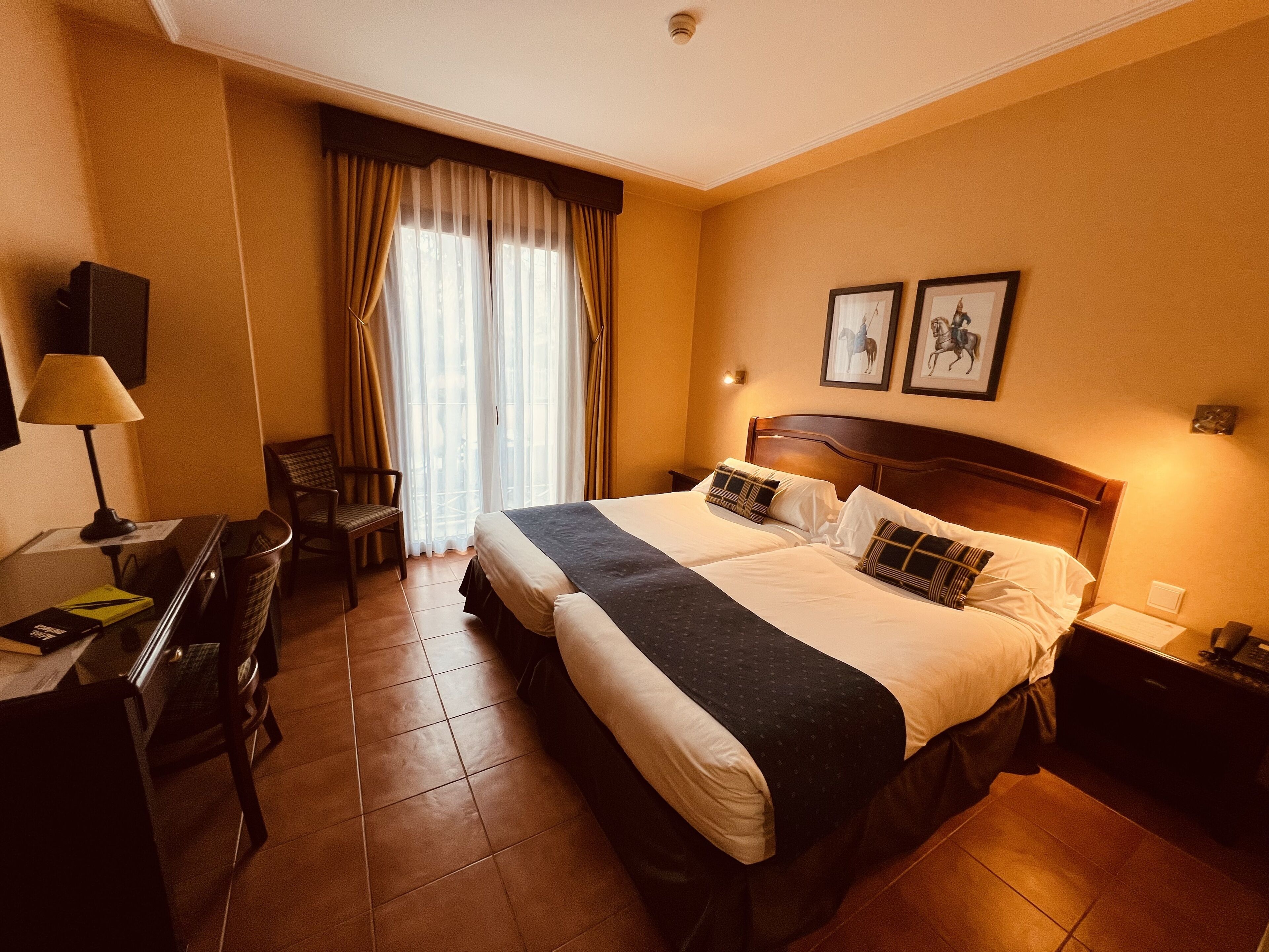 A hotel room with a large bed, a desk with a lamp, a chair, and a window with curtains.