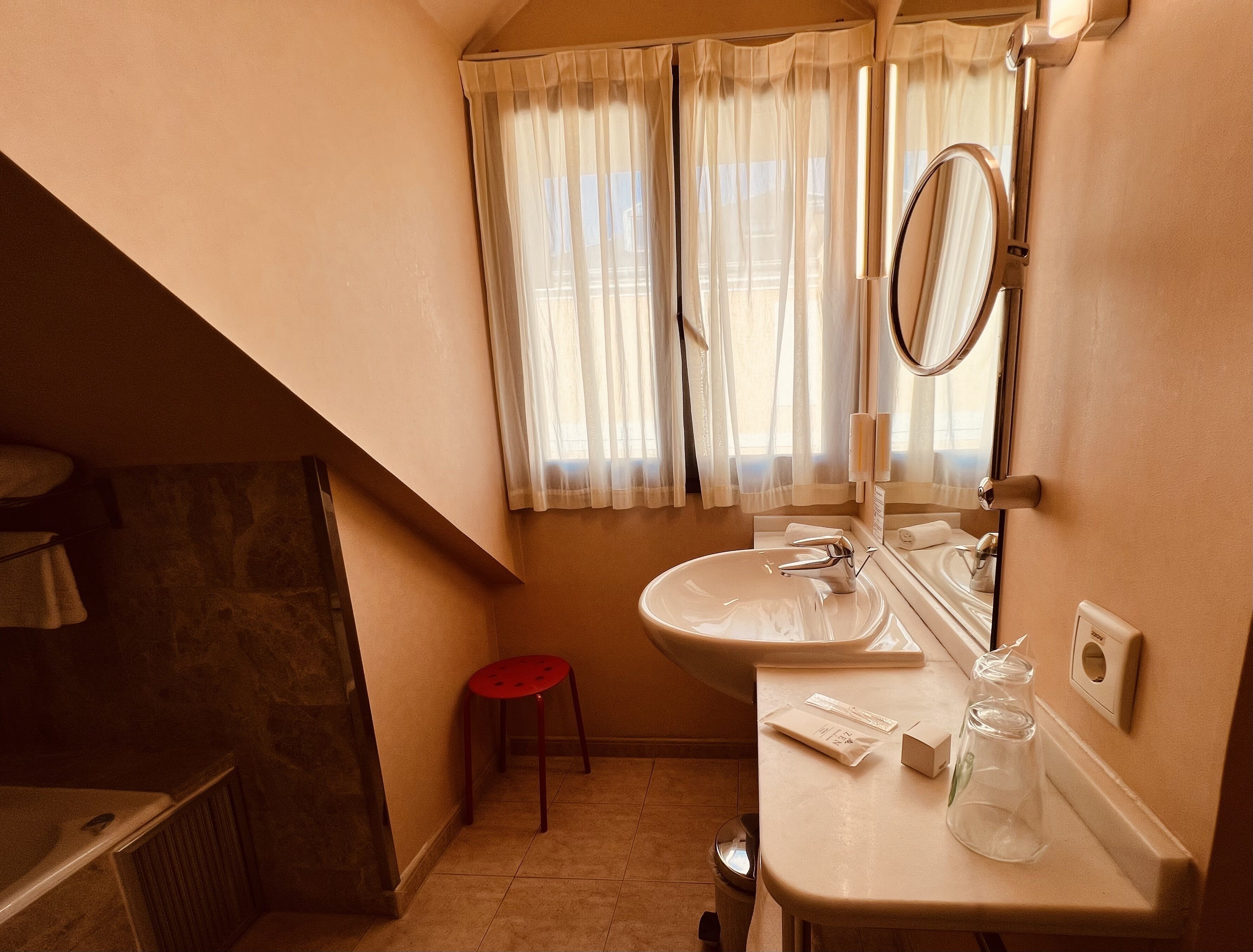Baño con lavamanos, espejo y ventana con cortinas. Se observa un taburete rojo y un perchero.