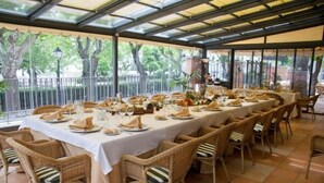 Una larga mesa de comedor preparada para una comida en un espacio al aire libre cubierto, con sillas de mimbre.