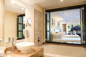 A modern bathroom with a large sink, bathtub, and a large mirror reflecting a bedroom with a bed and a view of the outdoors.