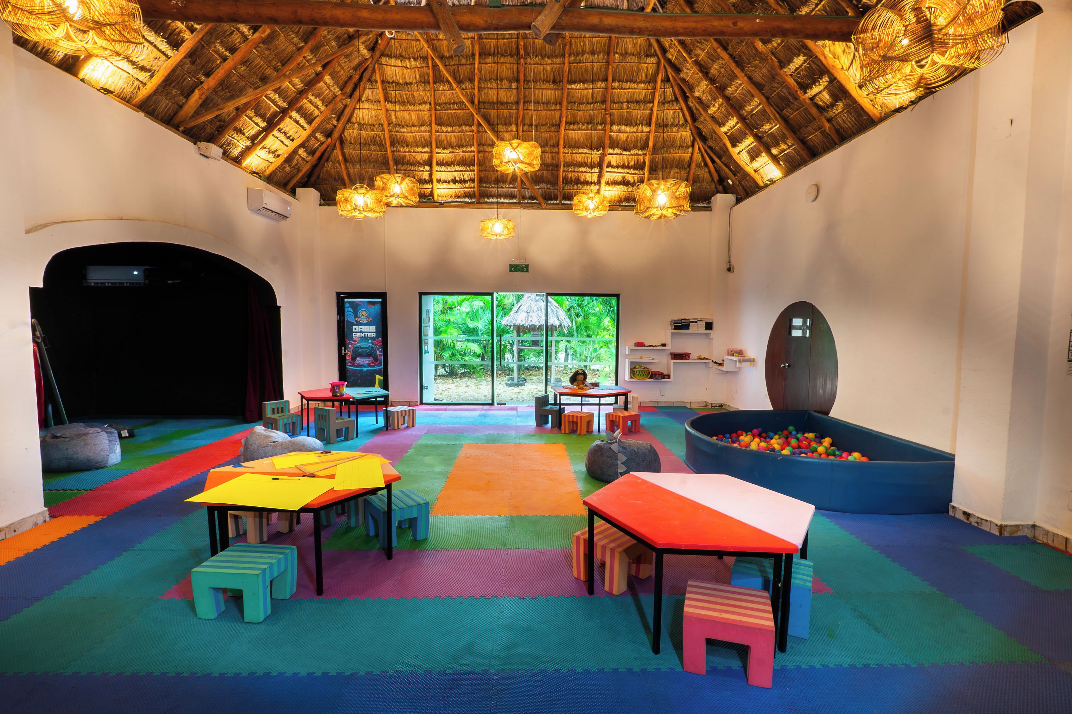 A room with a thatched roof, colorful play mats, tables, and a play area with a ball pit.