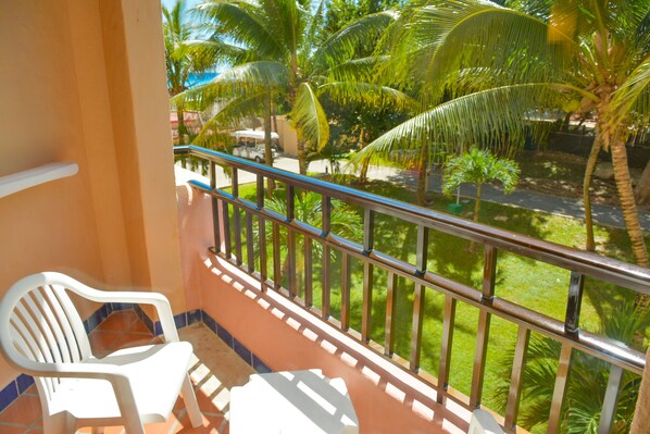 A balcony with a white plastic chair, a metal railing, and a view of palm trees and a road.
