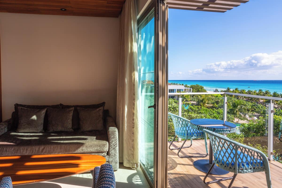 A balcony with a sofa, a table, and chairs, overlooking a turquoise ocean and greenery.