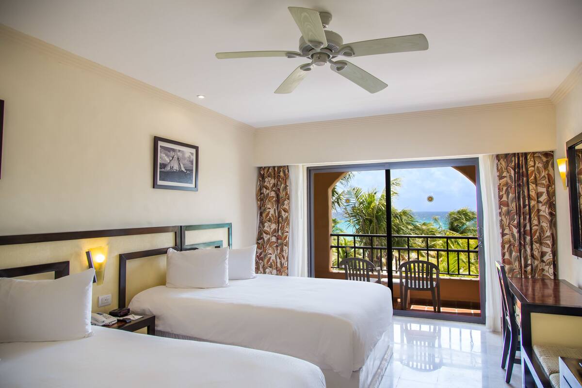 Une chambre d’hôtel avec deux lits, un ventilateur de plafond, un balcon avec vue sur l’océan et un coin repas.