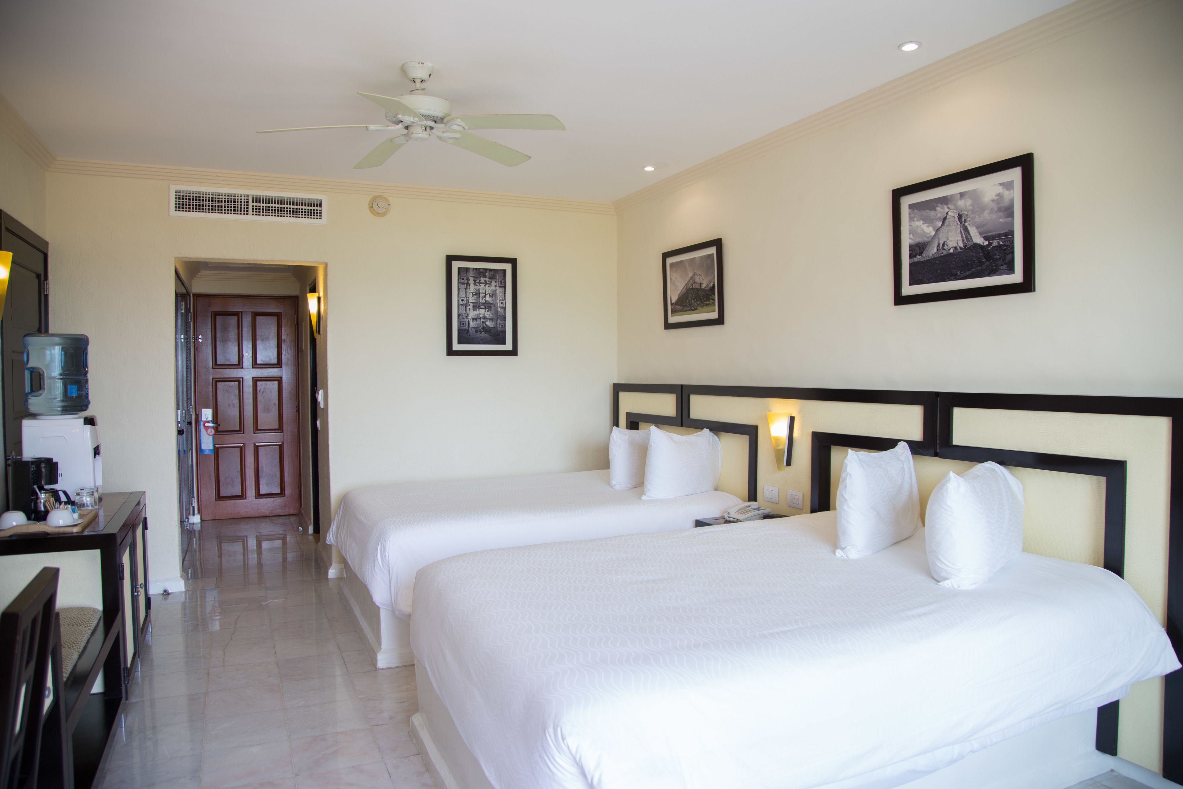 A hotel room with two beds, a desk with a chair, a ceiling fan, and framed pictures on the wall.