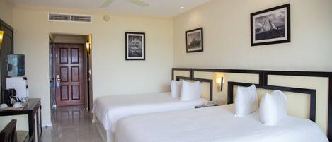Una habitación de hotel con dos camas, un escritorio con una silla, un ventilador de techo y cuadros en la pared.