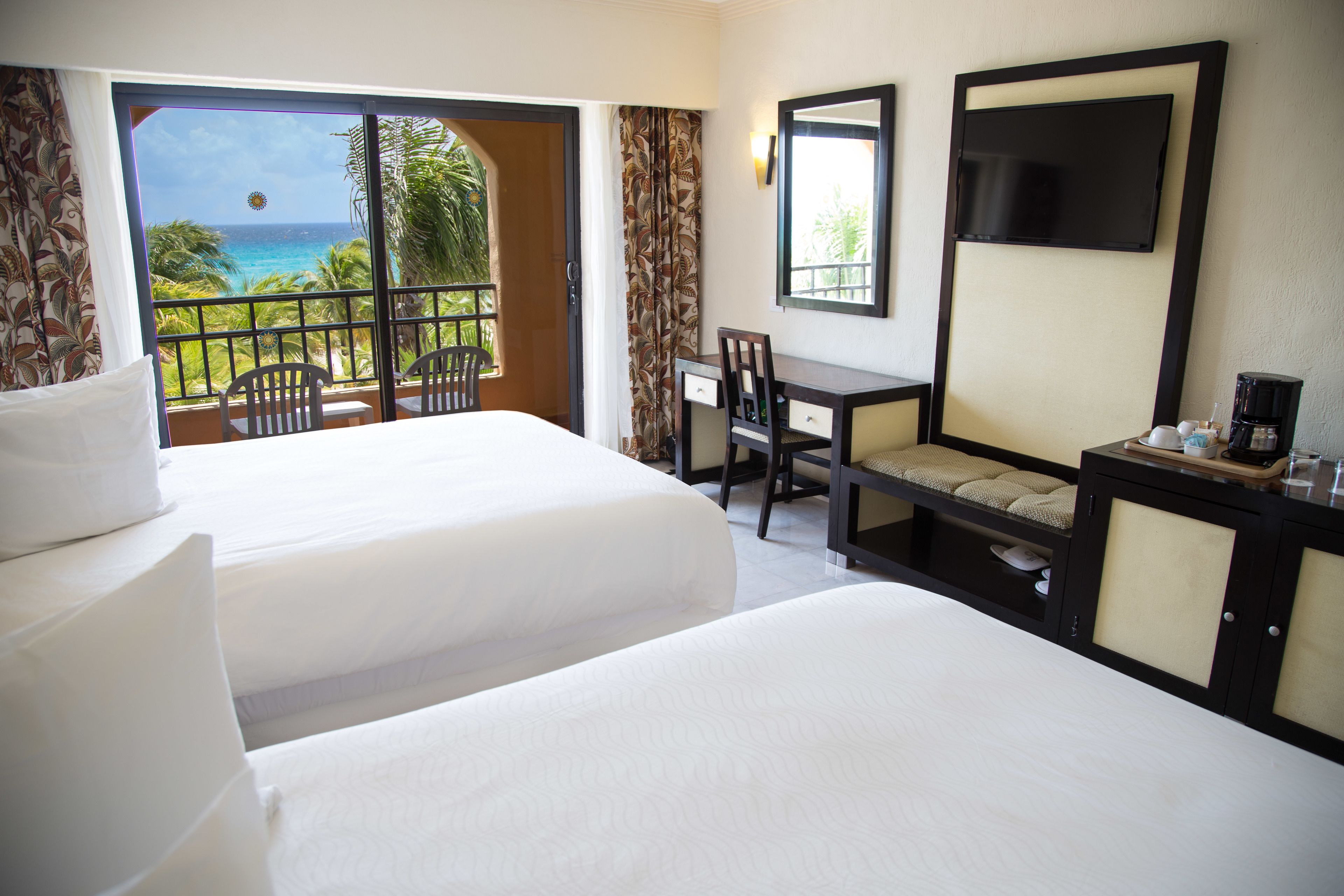 A hotel room with two beds, a desk, a chair, a TV, and a view of the ocean.