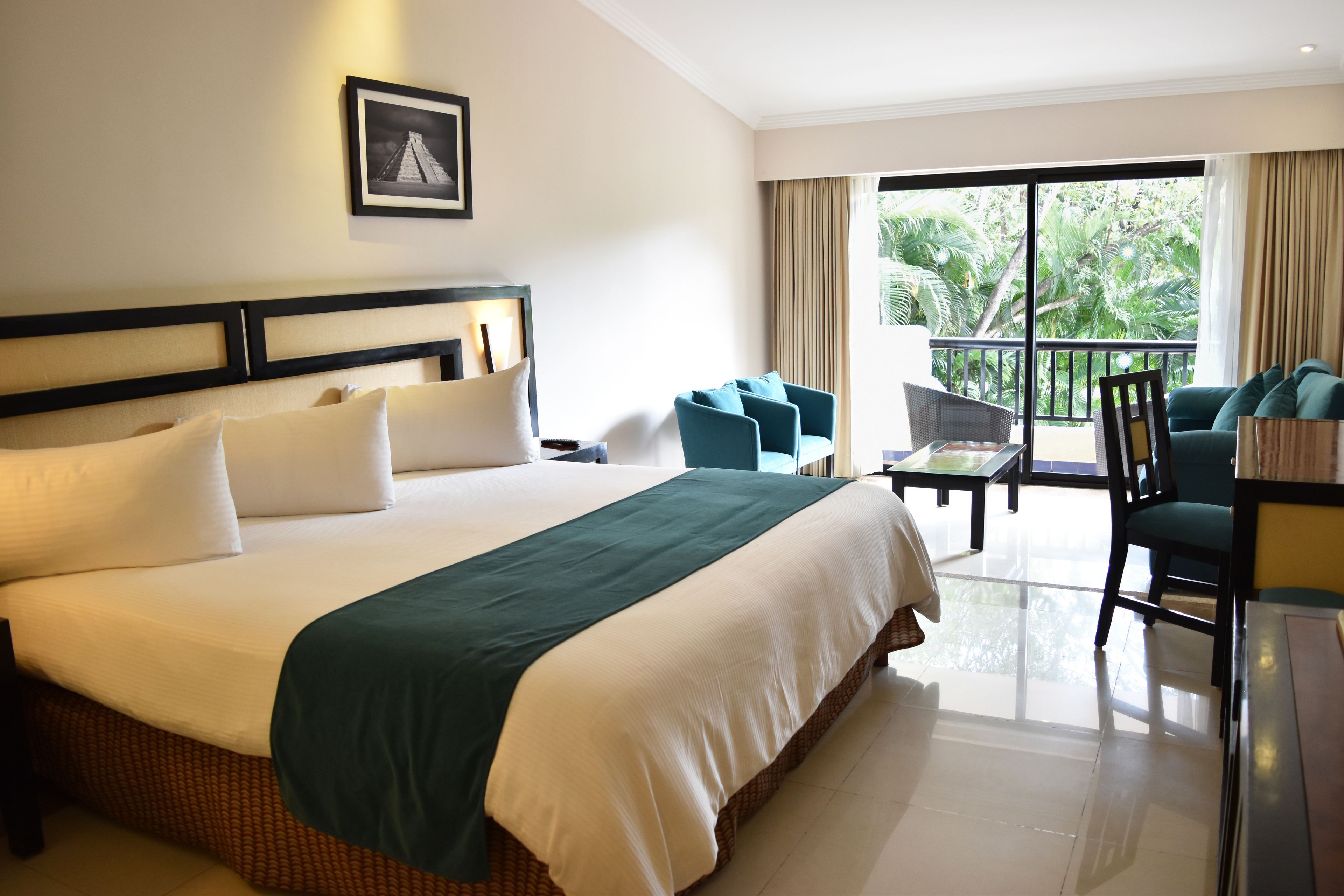 A hotel room with a large bed, a balcony with seating, and a view of greenery.