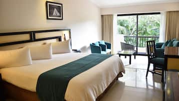A hotel room with a large bed, a balcony with seating, and a view of greenery.