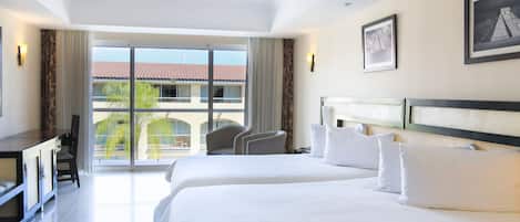 A hotel room with two beds, a desk, a chair, and a view of a building with a red roof through a large window.