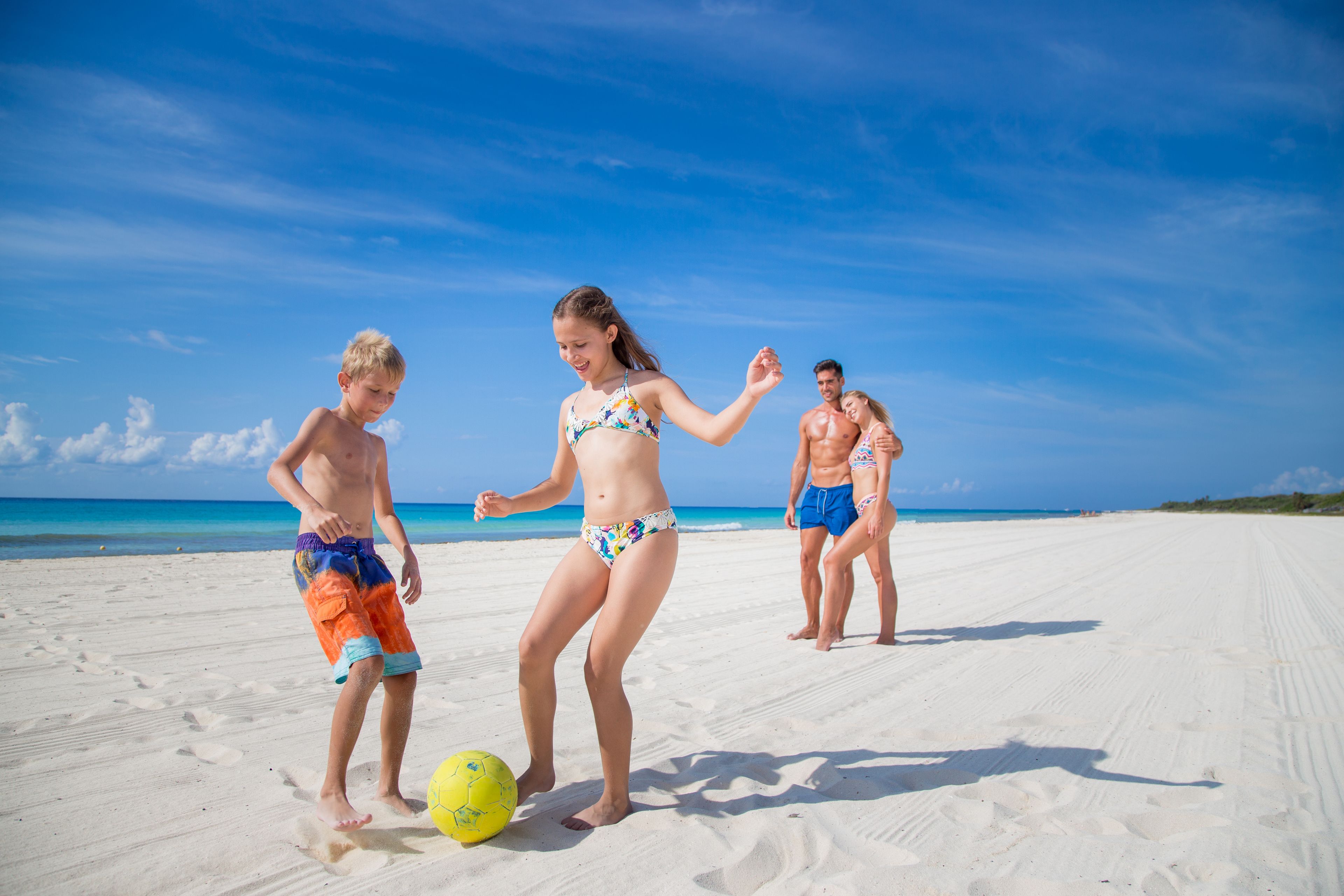 A beach scene with three people playing with a yellow ball.