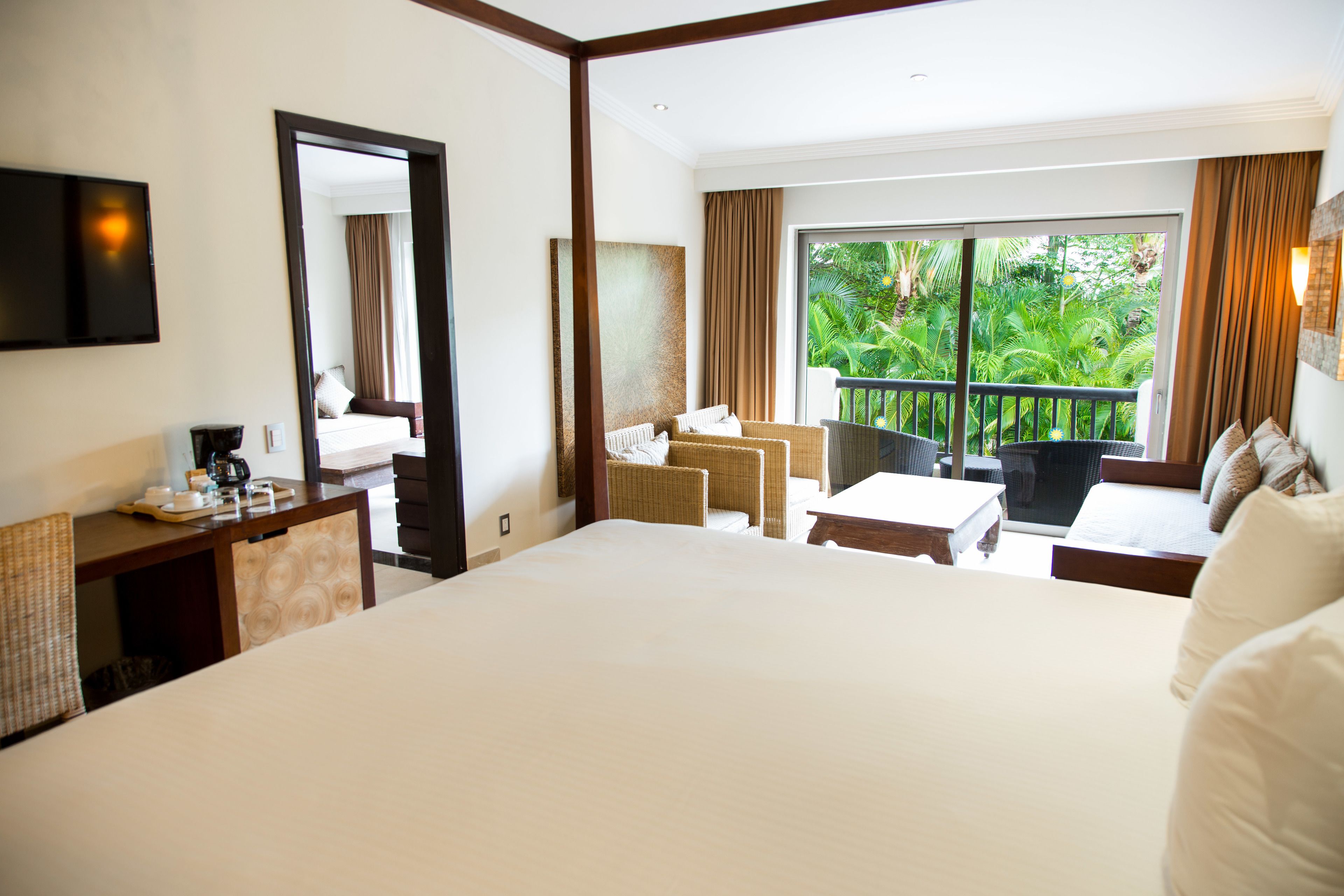 A hotel room with a large bed, a desk with a coffee maker, a seating area with chairs, and a balcony with a view of greenery.