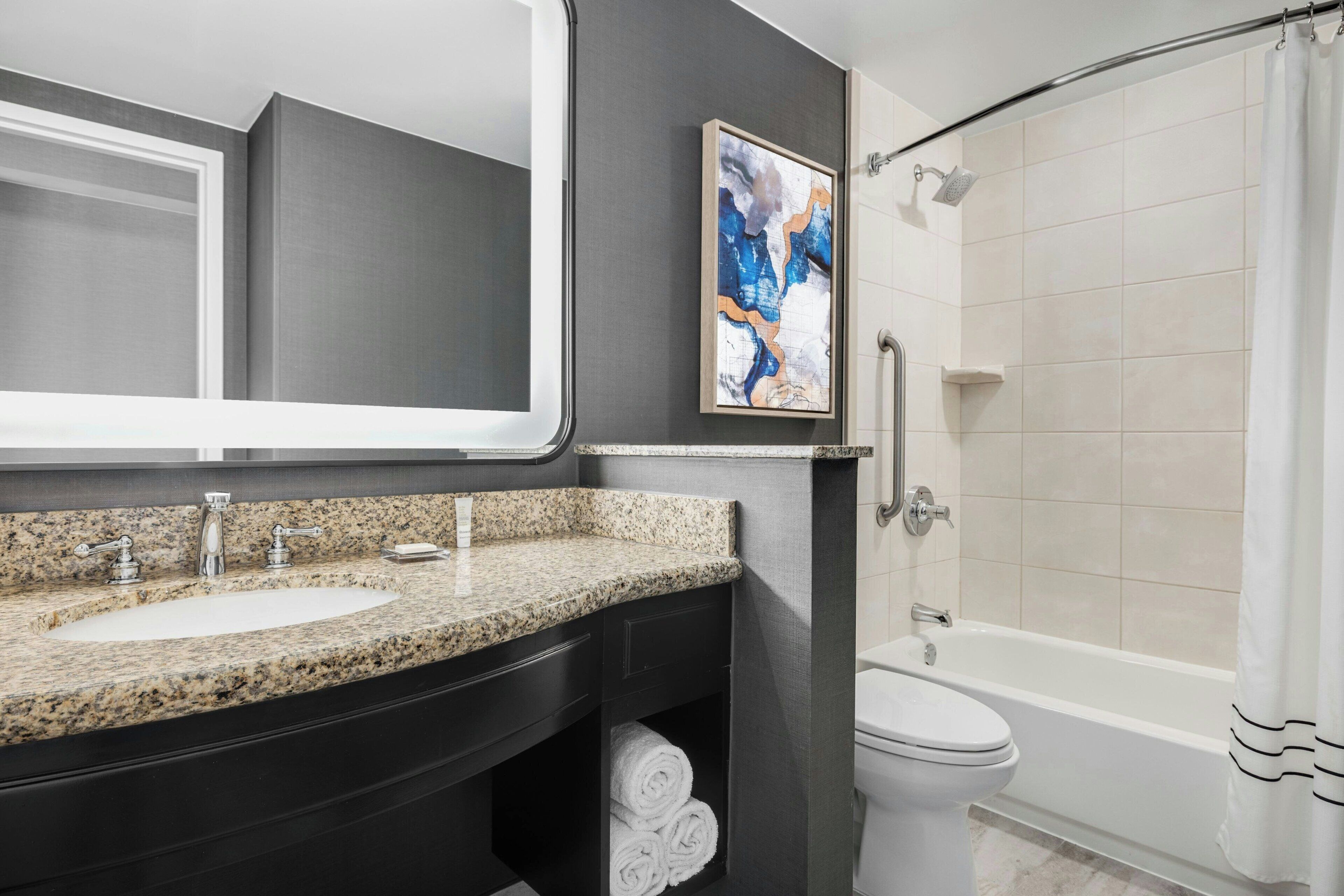A modern bathroom with a granite countertop sink, a large mirror, a framed abstract painting, a white toilet, and a walk-in shower with a white curtain.