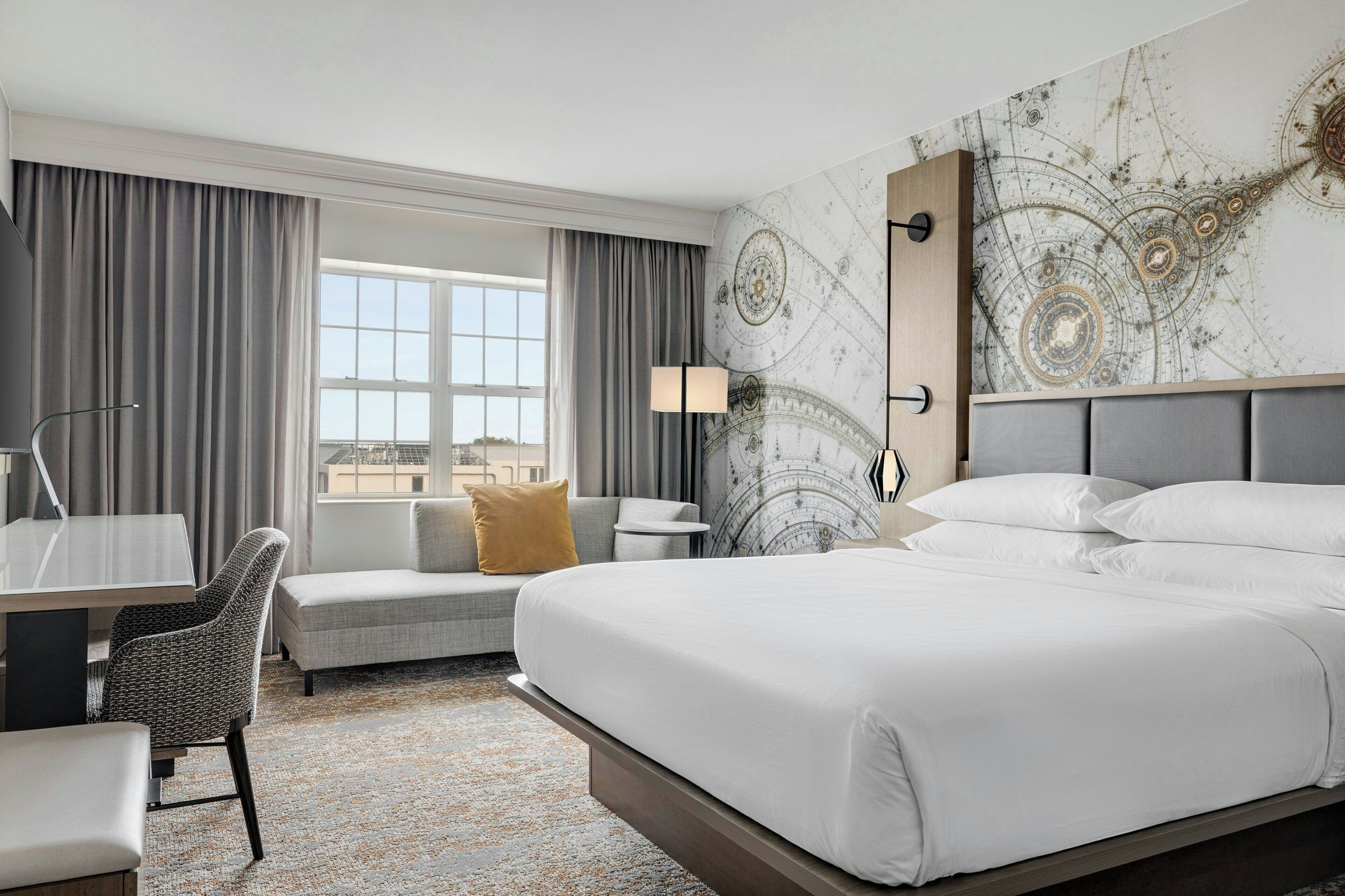 A hotel room with a large bed, a desk with a chair, a sofa, and a large window with curtains.
