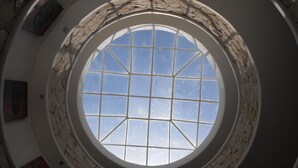 Een rond dakvenster met een rasterpatroon, omringd door stenen muren en omkadert kunstwerken.