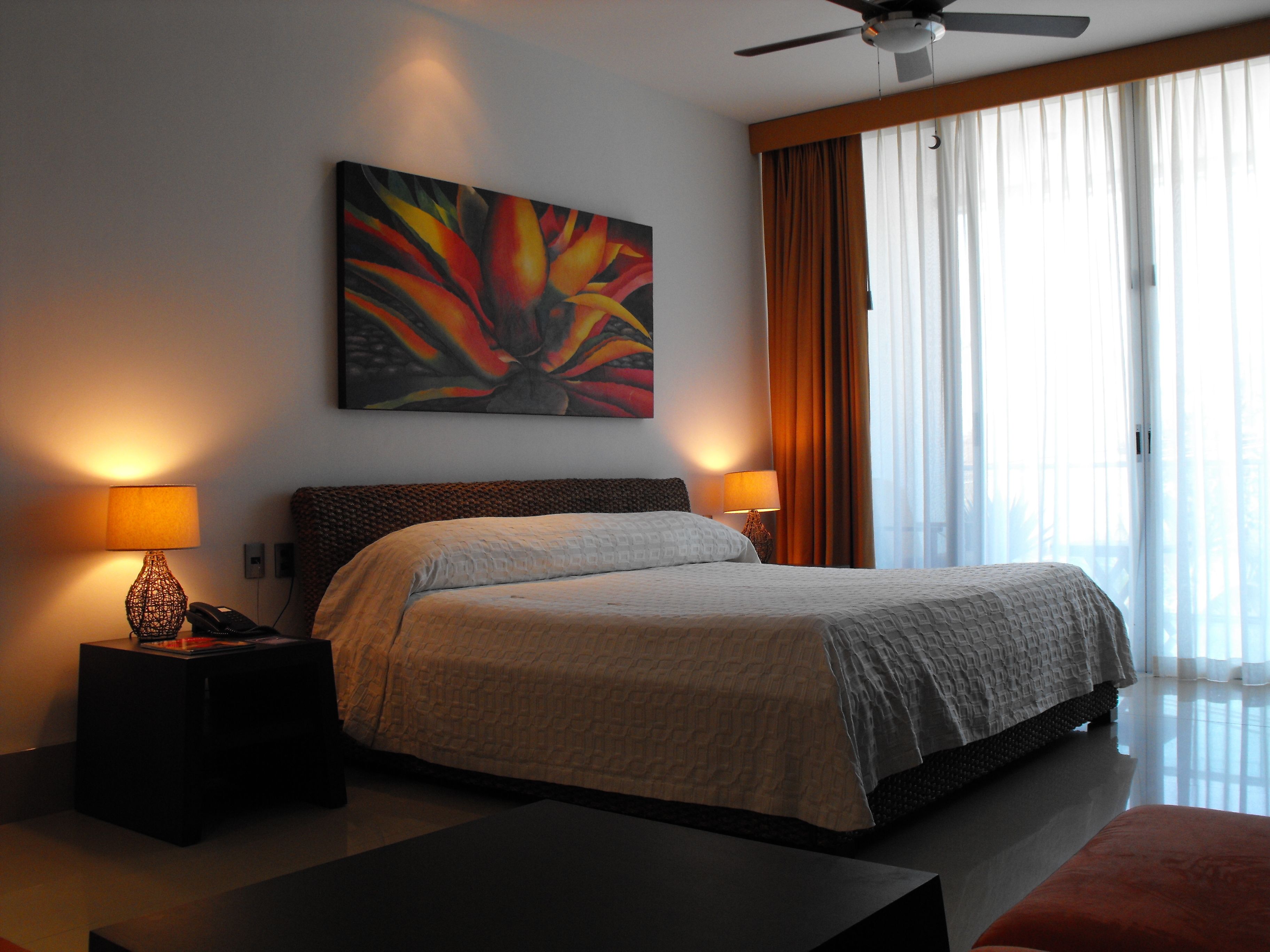 A hotel room with a bed, two bedside lamps, a ceiling fan, a painting on the wall, and a window with curtains.