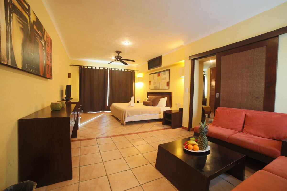 Ein Hotelzimmer mit einem Bett, einem roten Sofa, einem Couchtisch mit Früchten, einem Fernseher und einem Deckenventilator.