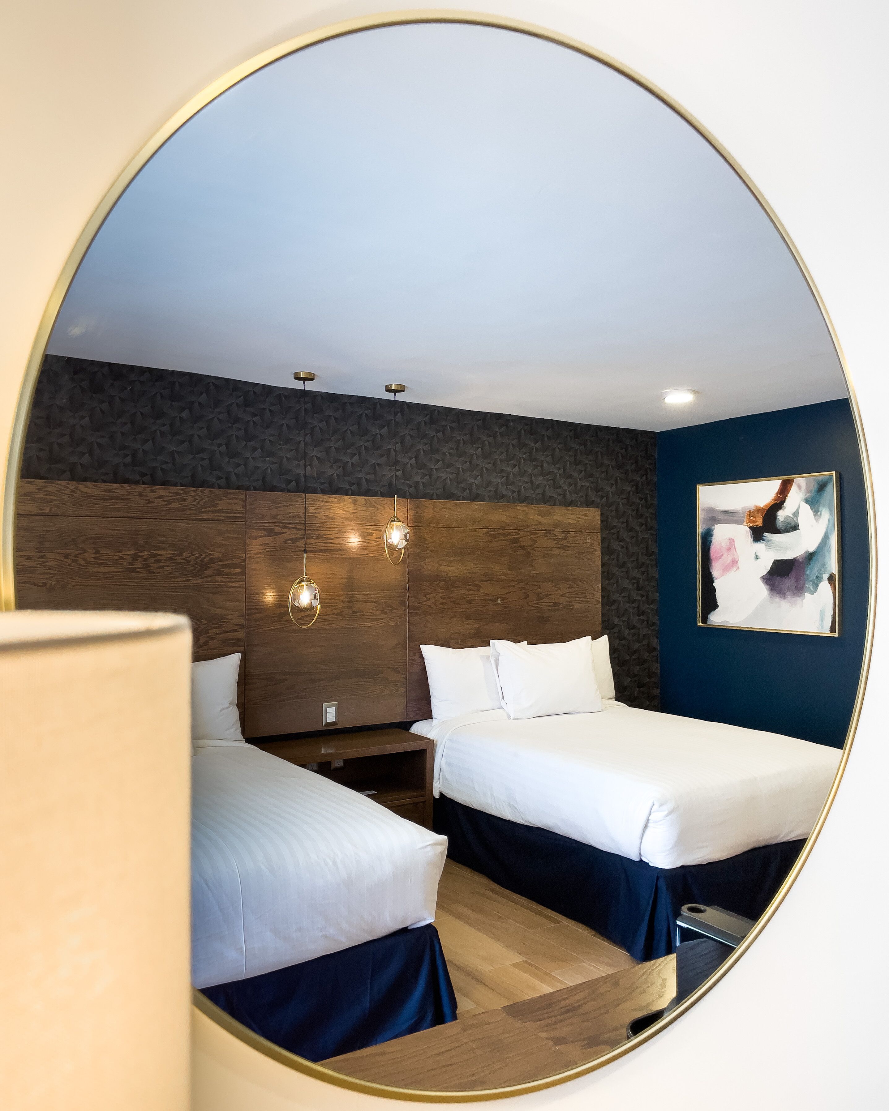A hotel room with two beds, a wooden headboard, a dark textured wall, a painting, and a round mirror.