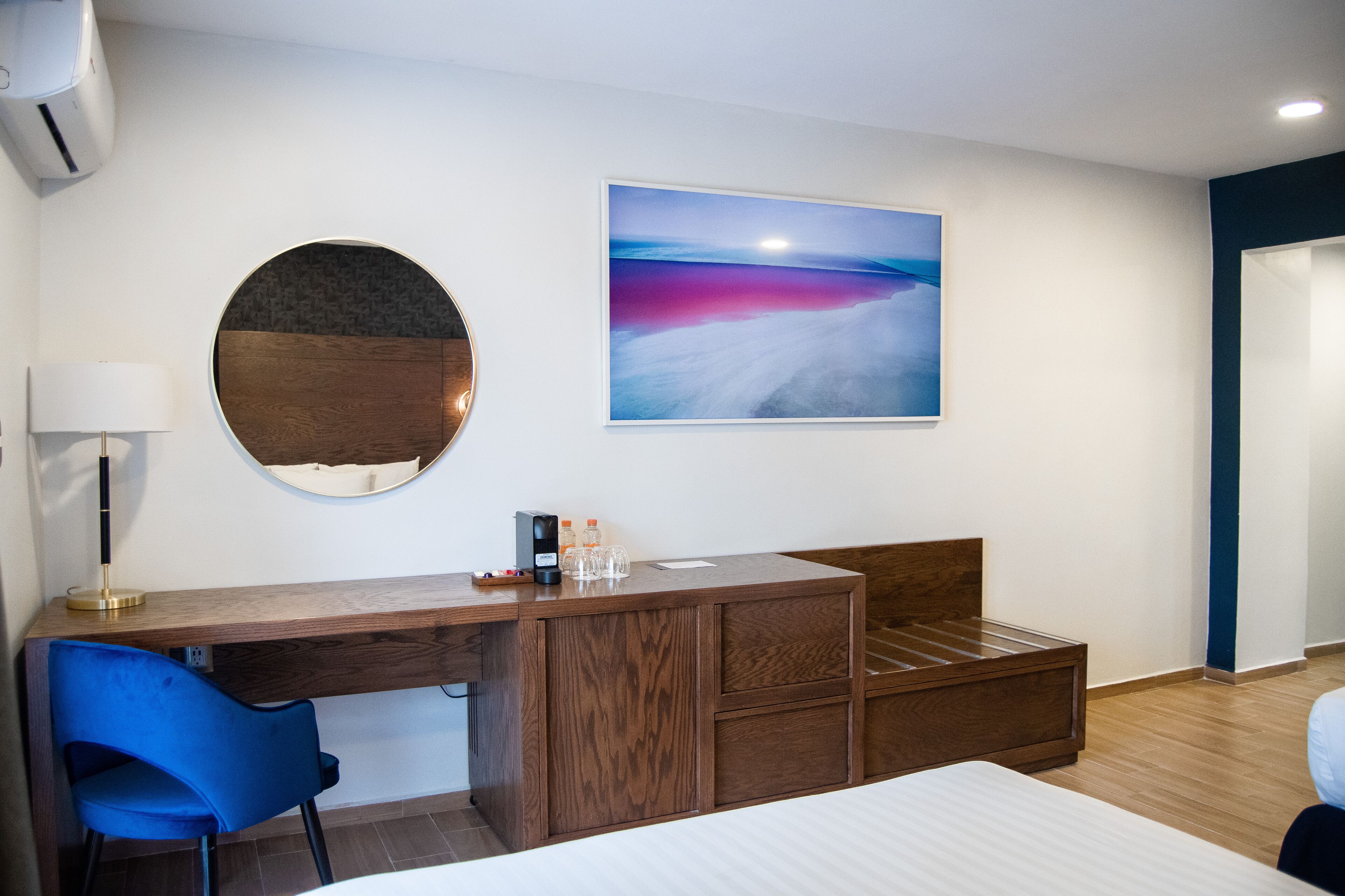 Habitación de hotel con un escritorio de madera, una silla azul, una cama, un espejo redondo y un cuadro de un atardecer sobre el agua.