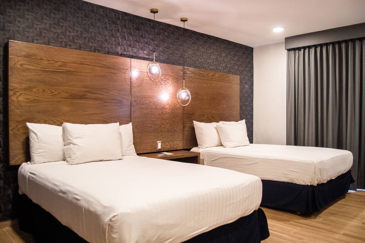 Une chambre d’hôtel avec deux lits, une tête de lit en bois, un mur orné d’un motif géométrique et une grande fenêtre avec des rideaux.