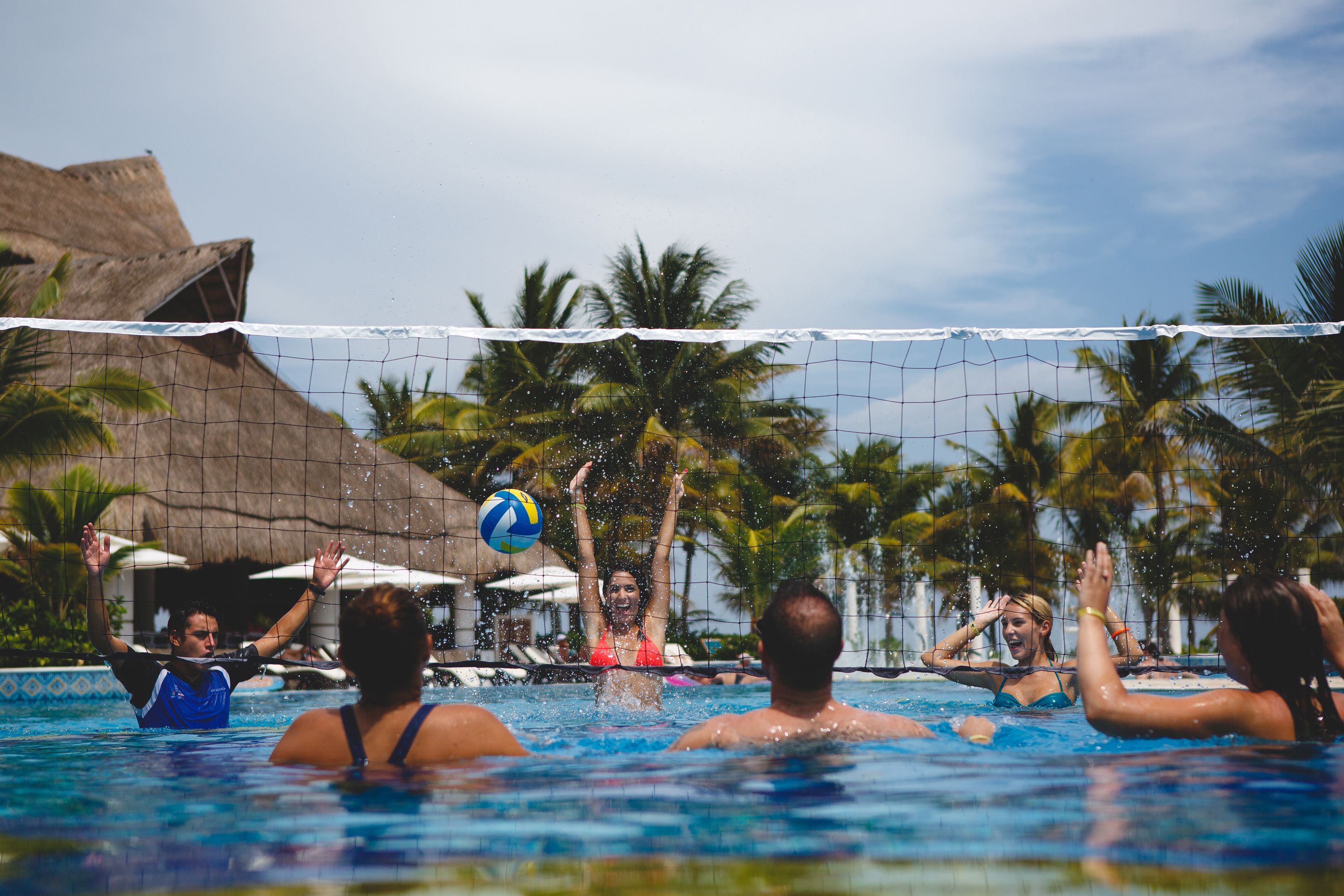A swimming pool with a volleyball net, palm trees, and a thatched-roof structure in the background.