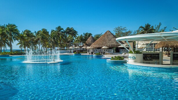 A swimming pool with a fountain, palm trees, and thatched-roof structures. - Catalonia Royal Tulum  Adults Only - All Inclusive (Xpu-Ha)