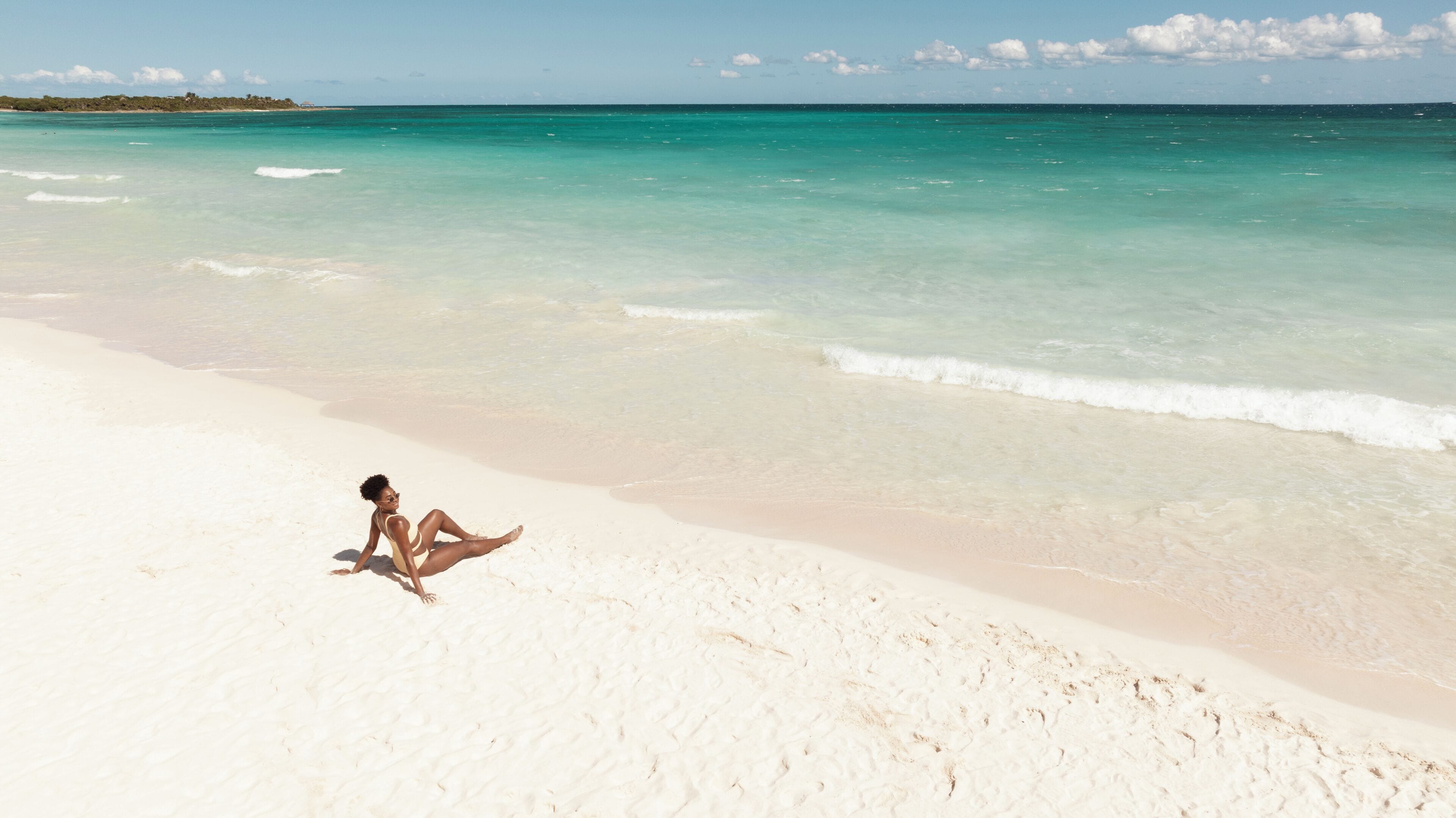 Una persona sentada en una playa de arena con agua turquesa cristalina y una costa distante.