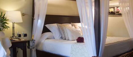 A four-poster bed with white linens and a canopy, a bedside table with a lamp, and a small potted plant.