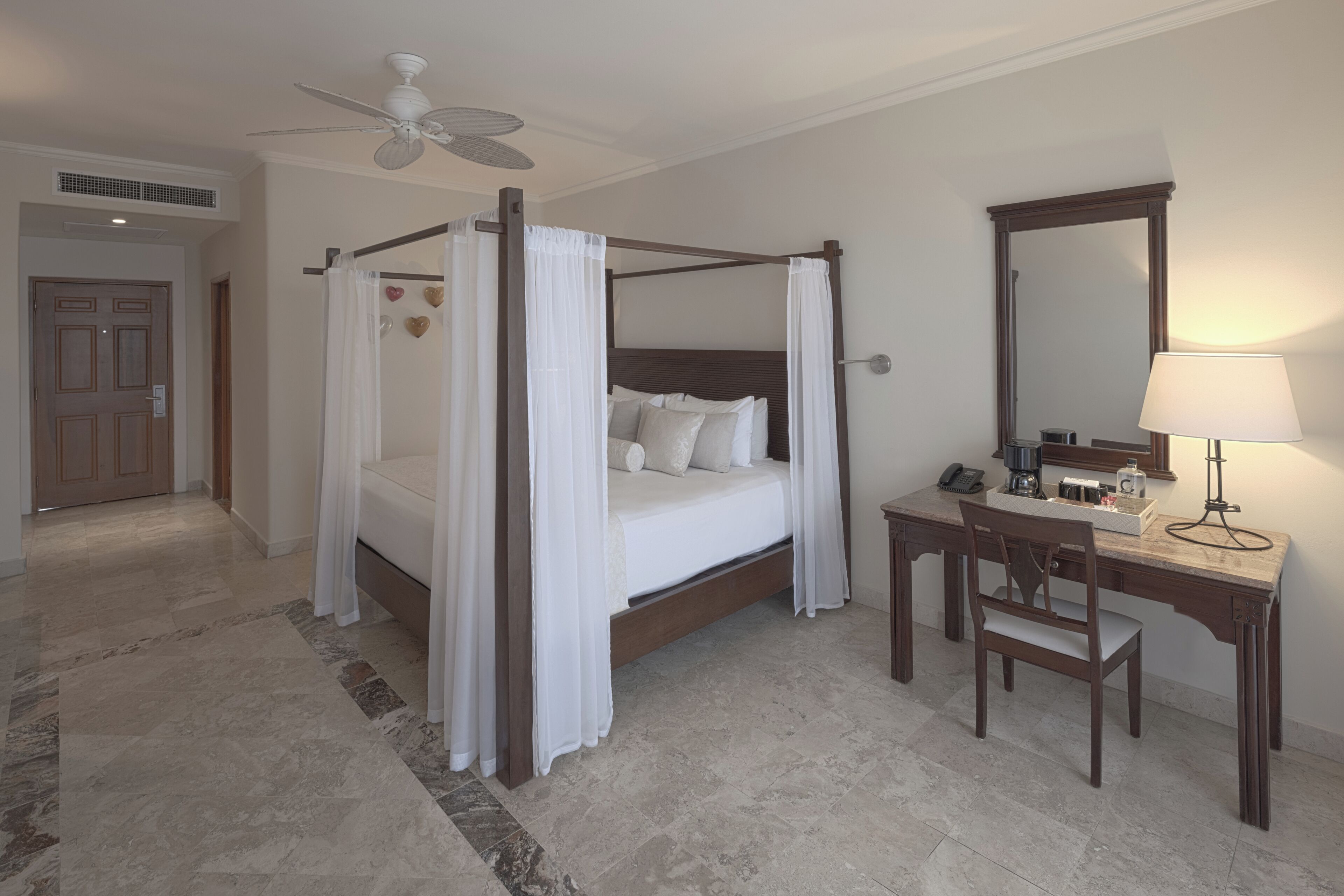 A four-poster bed with white linens, a wooden desk with a lamp, and a mirror.