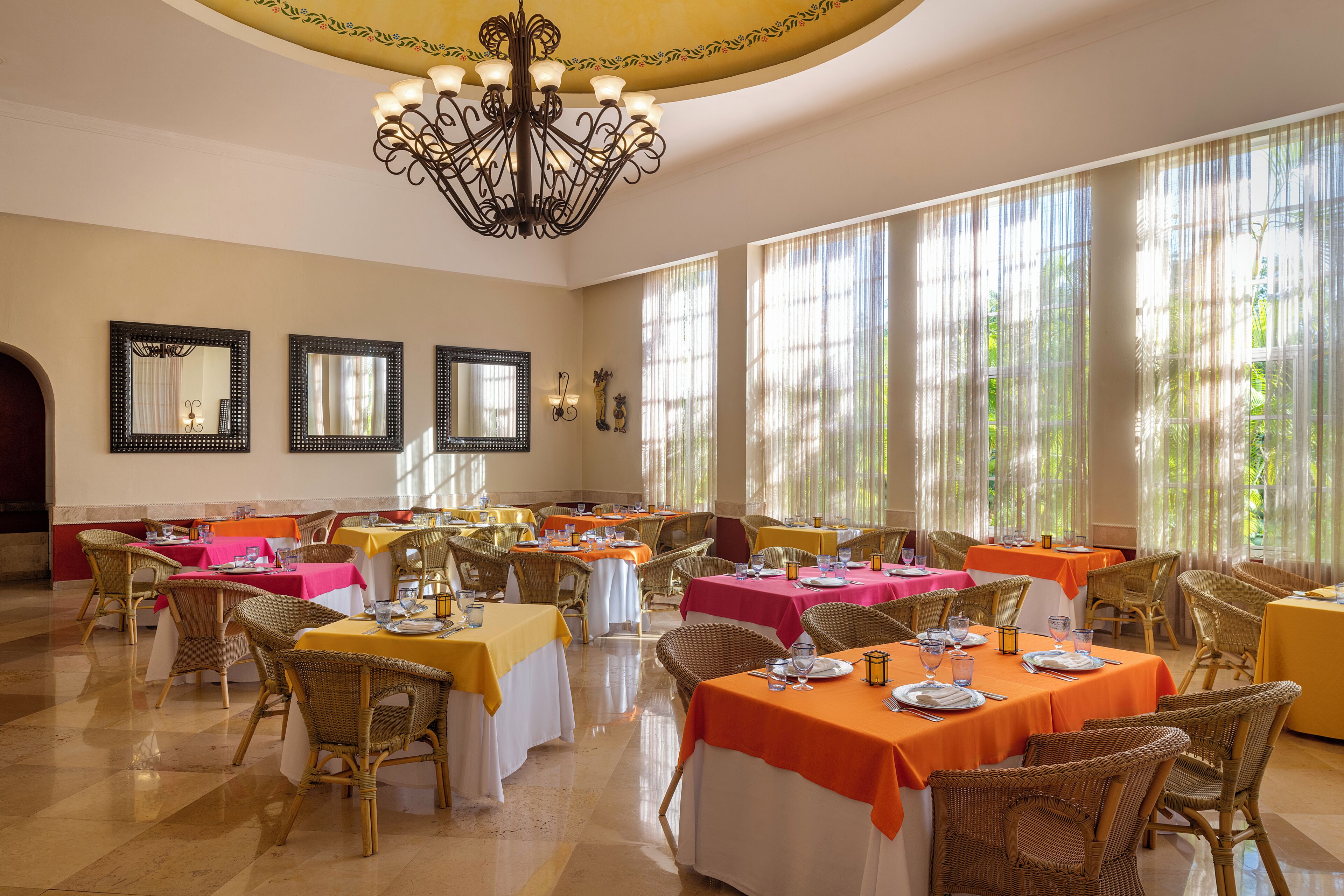 A dining area with tables set for multiple guests, wicker chairs, and a chandelier.