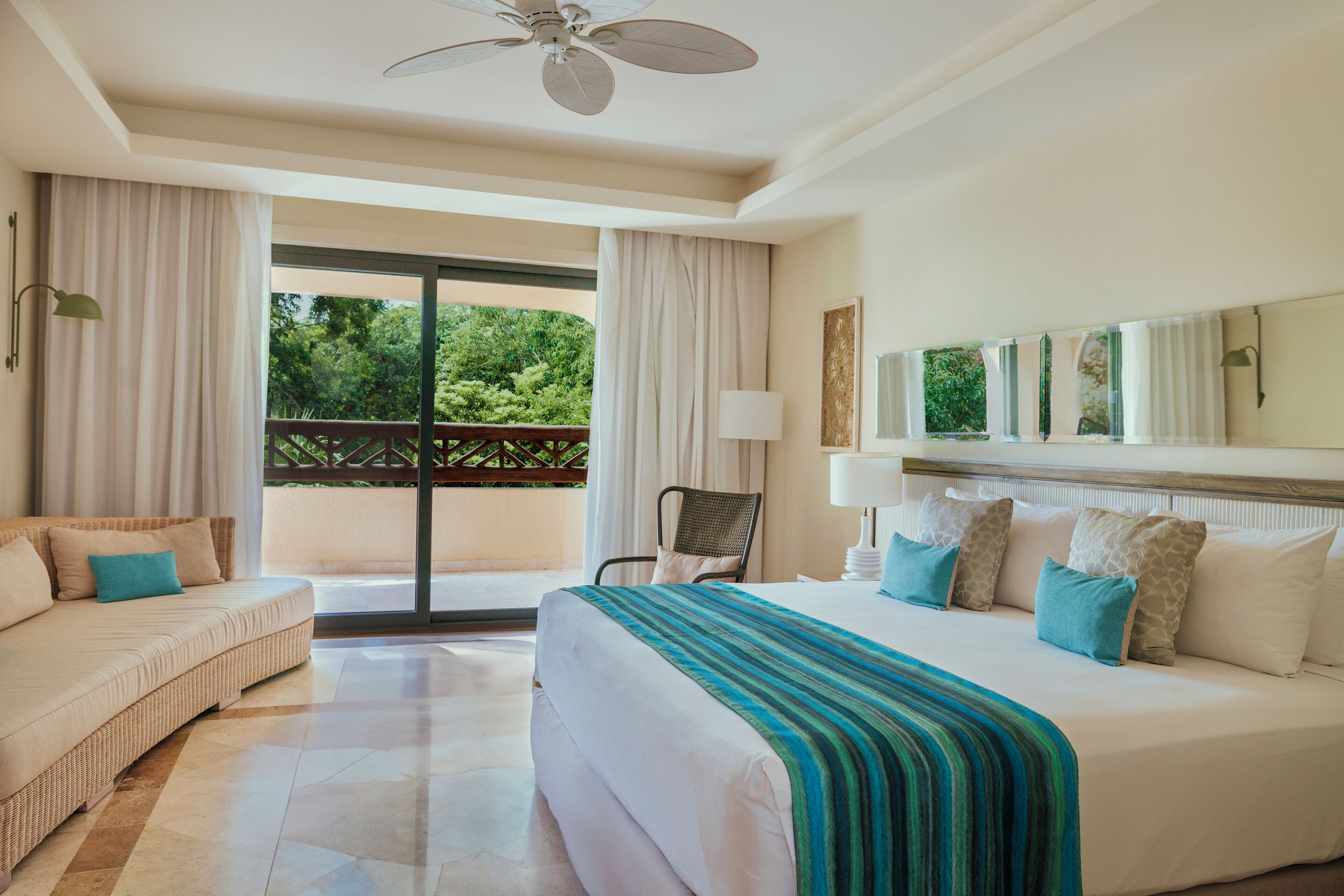 A hotel room with a bed, a sofa, a balcony with curtains, a ceiling fan, and a view of greenery.