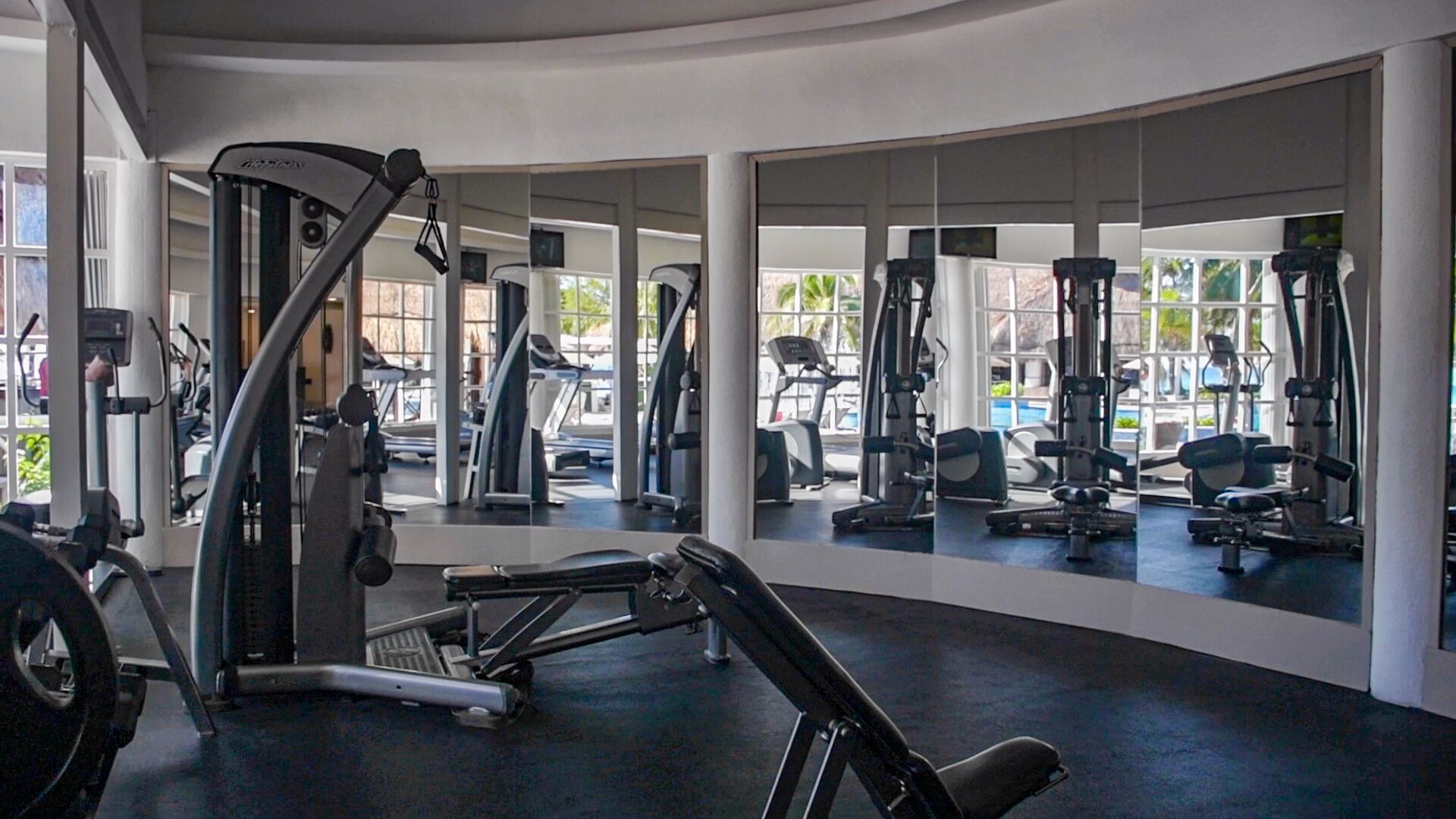 Un gimnasio con diversas máquinas de ejercicio, ventanas amplias y una distribución en forma de curva.