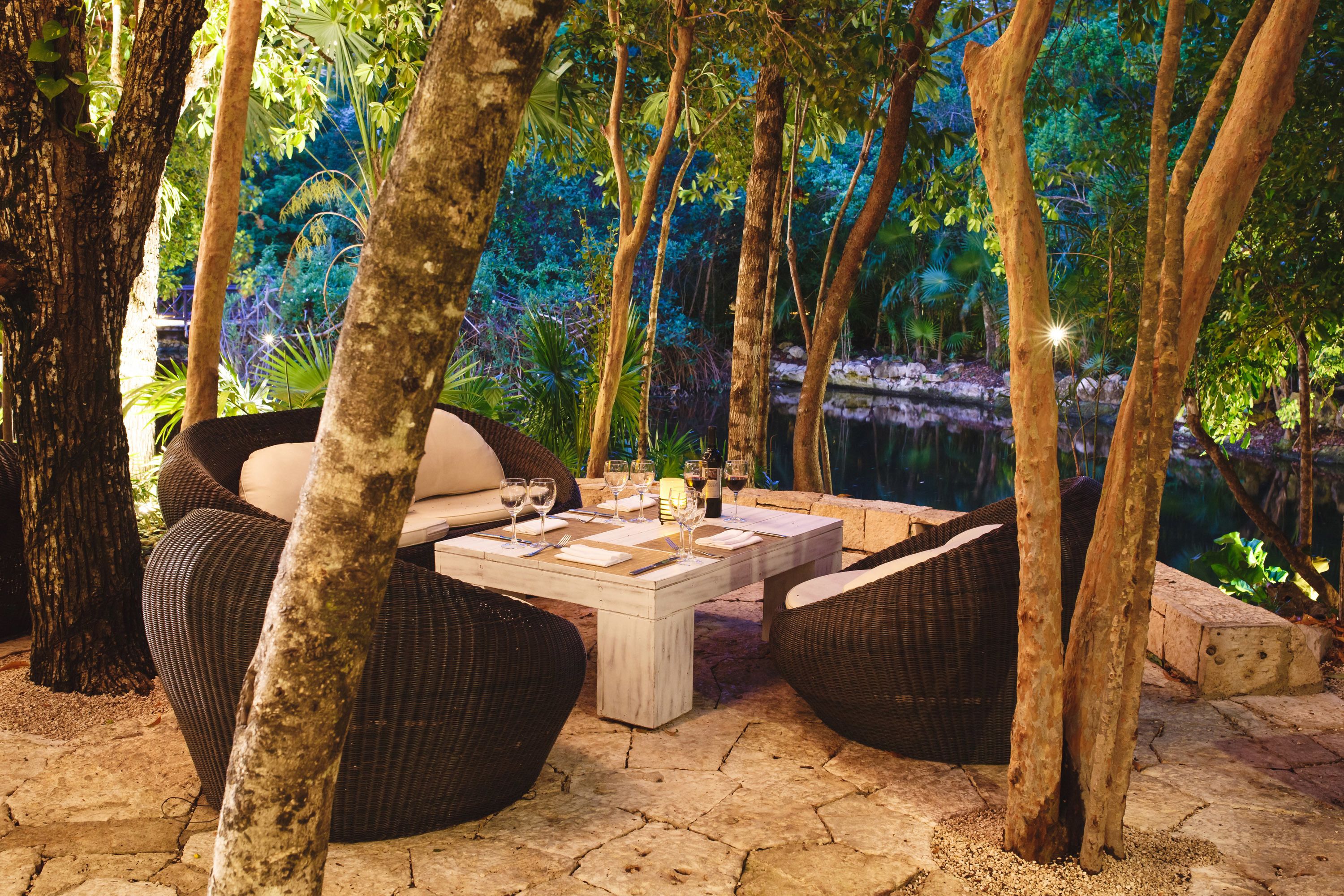 An outdoor dining area with wicker furniture and a stone table, surrounded by trees and a pond.