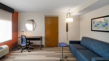 A hotel room with a wooden door, a desk with a chair, a blue sofa, a standing lamp, and a wall-mounted mirror.