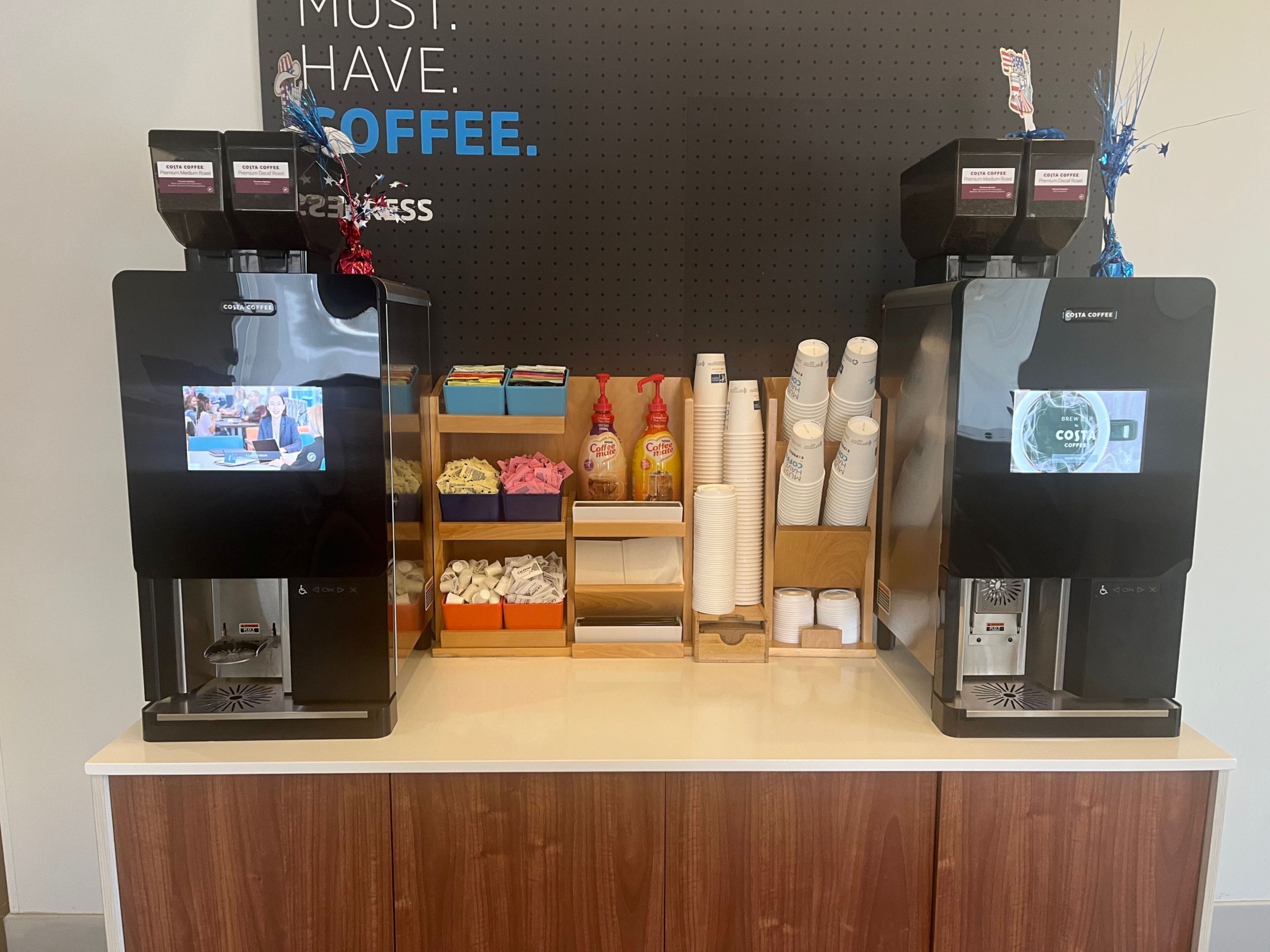 A coffee station with two coffee dispensers, a shelf with coffee pods, and a display of disposable cups.