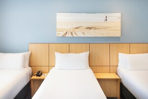 A hotel room with two beds, a wooden headboard, a nightstand with a phone, and a painting of a beach scene.
