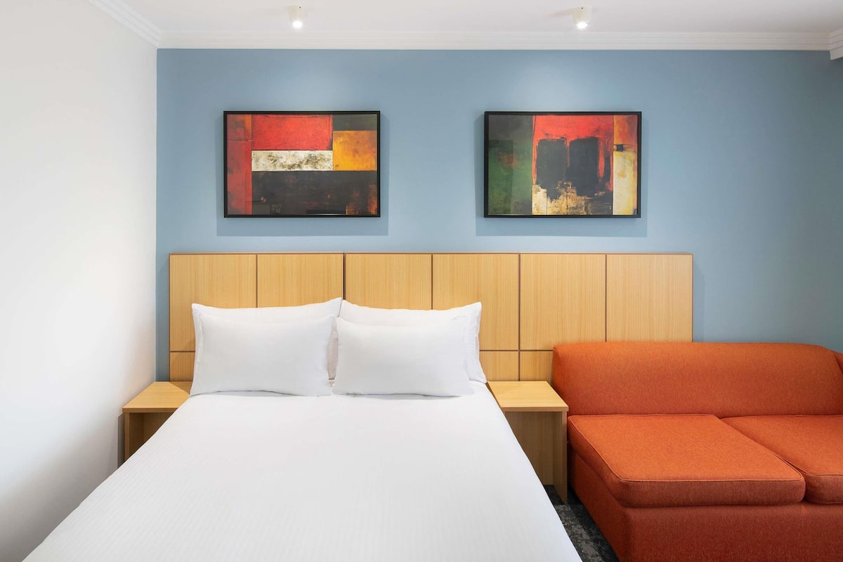Een hotelkamer met een bed, twee nachtkastjes, een oranje sofa en twee abstracte schilderijen aan de muur.