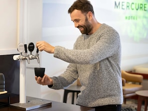 Une personne manipule un robinet à deux becs portant les inscriptions « PUREZZA SPARKLING » et « PUREZZA STILL », dans un décor moderne, avec une enseigne « MERCURE HOTEL » en arrière-plan.