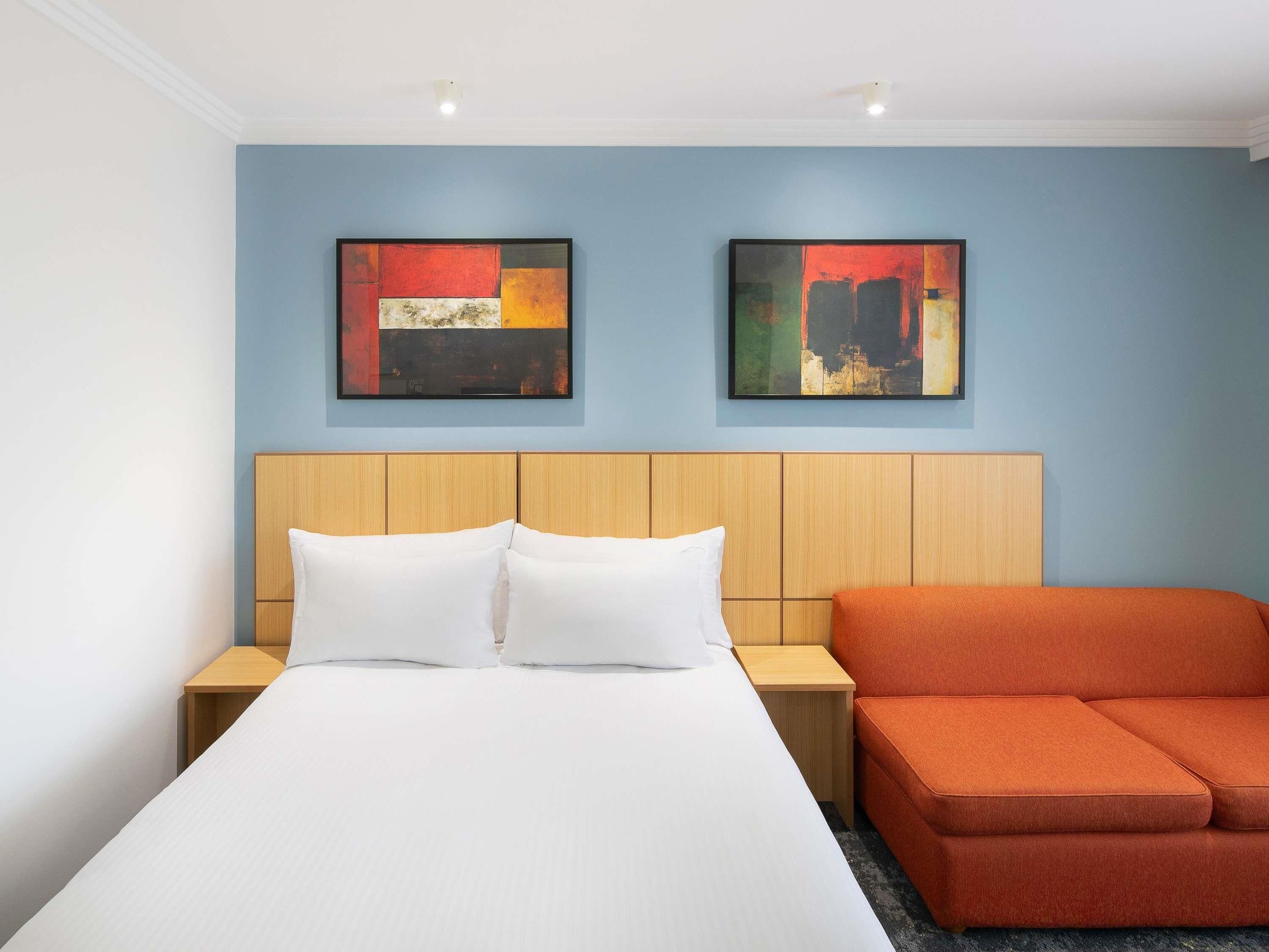 A hotel room with a bed, two nightstands, an orange sofa, and two abstract paintings on the wall.
