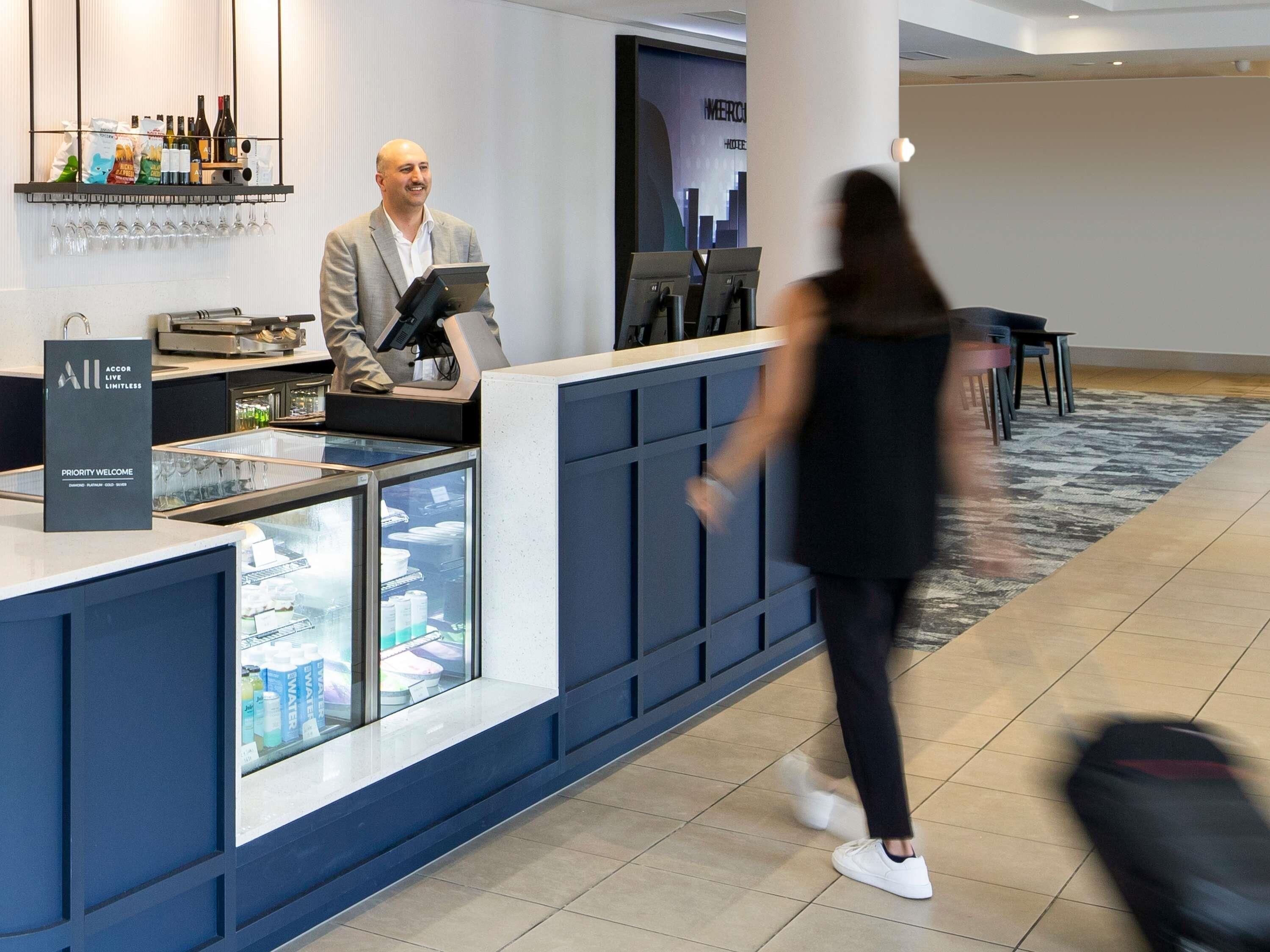 A hotel reception desk with a man behind it, a glass display case with bottled water, and a sign that reads 'Priority Welcome.'