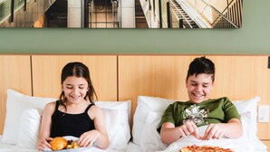 Twee personen zitten op een bed met kussens, één met een bord eten en de ander met een pizza.