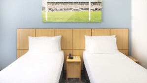 Ein Hotelzimmer mit zwei Betten, einem Holz-Kopfteil, einem kleinen Nachttisch mit Telefon und Fernbedienung sowie einem gerahmten Bild eines Stadions an der Wand.