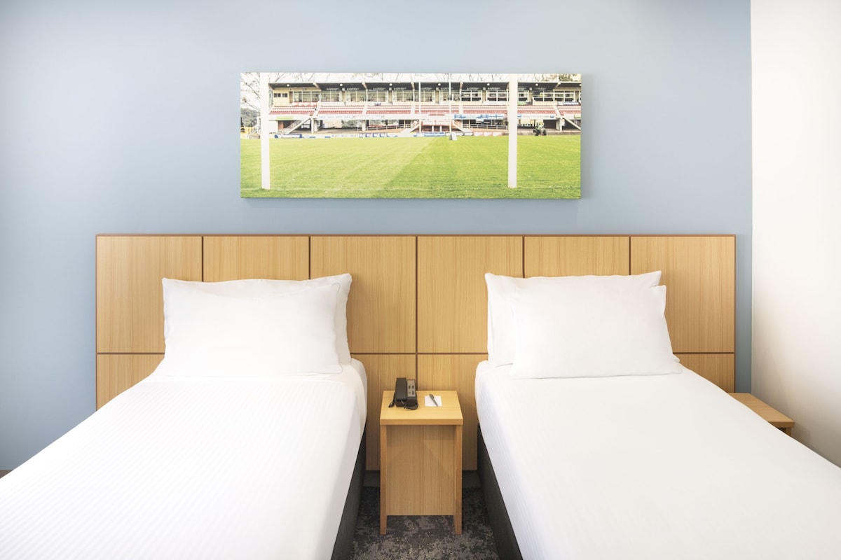 A hotel room with two beds, a wooden headboard, a small bedside table with a phone and a remote, and a framed picture of a stadium on the wall.