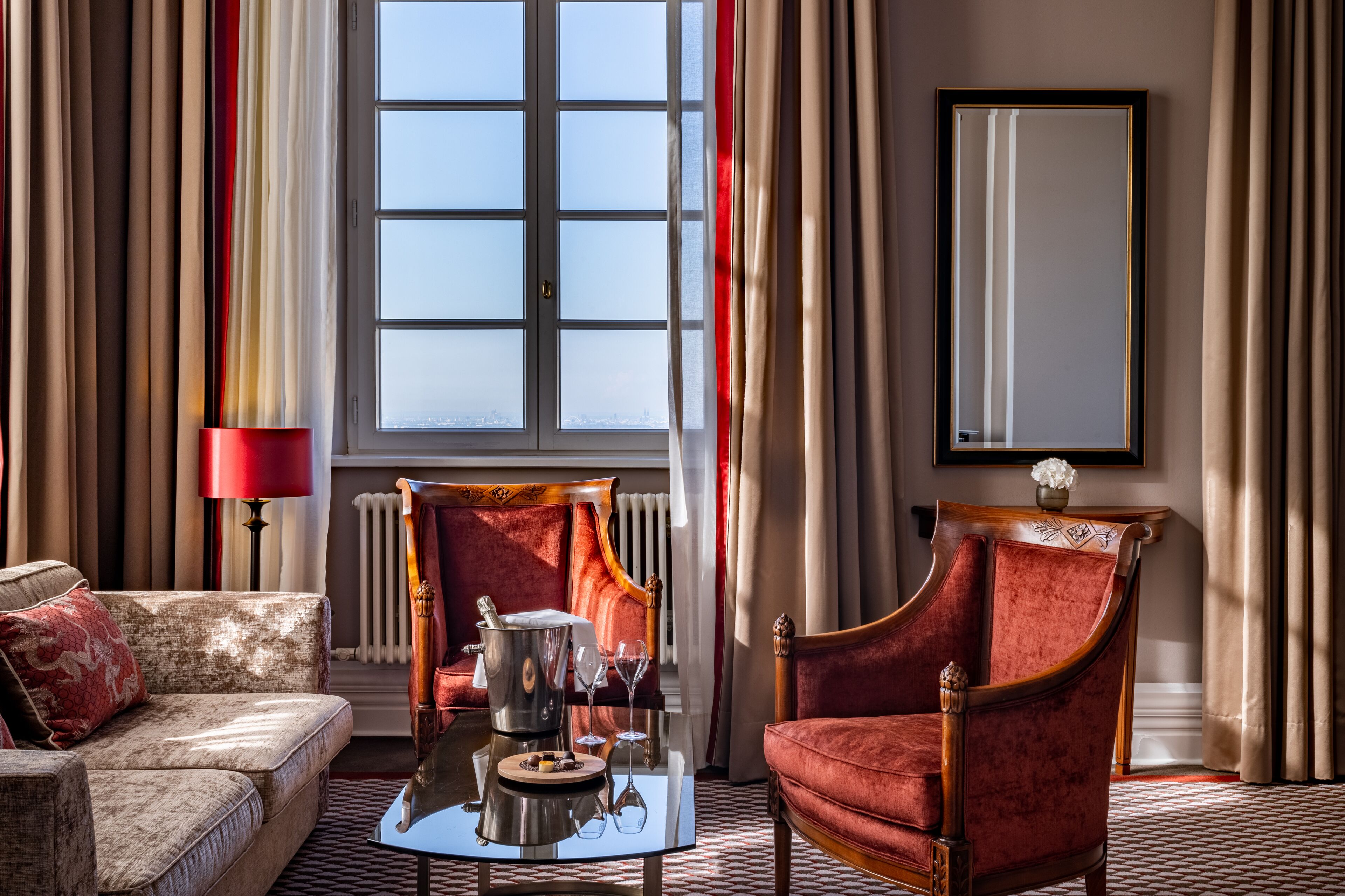 A room with a sofa, a red armchair, a table with a bottle and glasses, and a window with curtains.